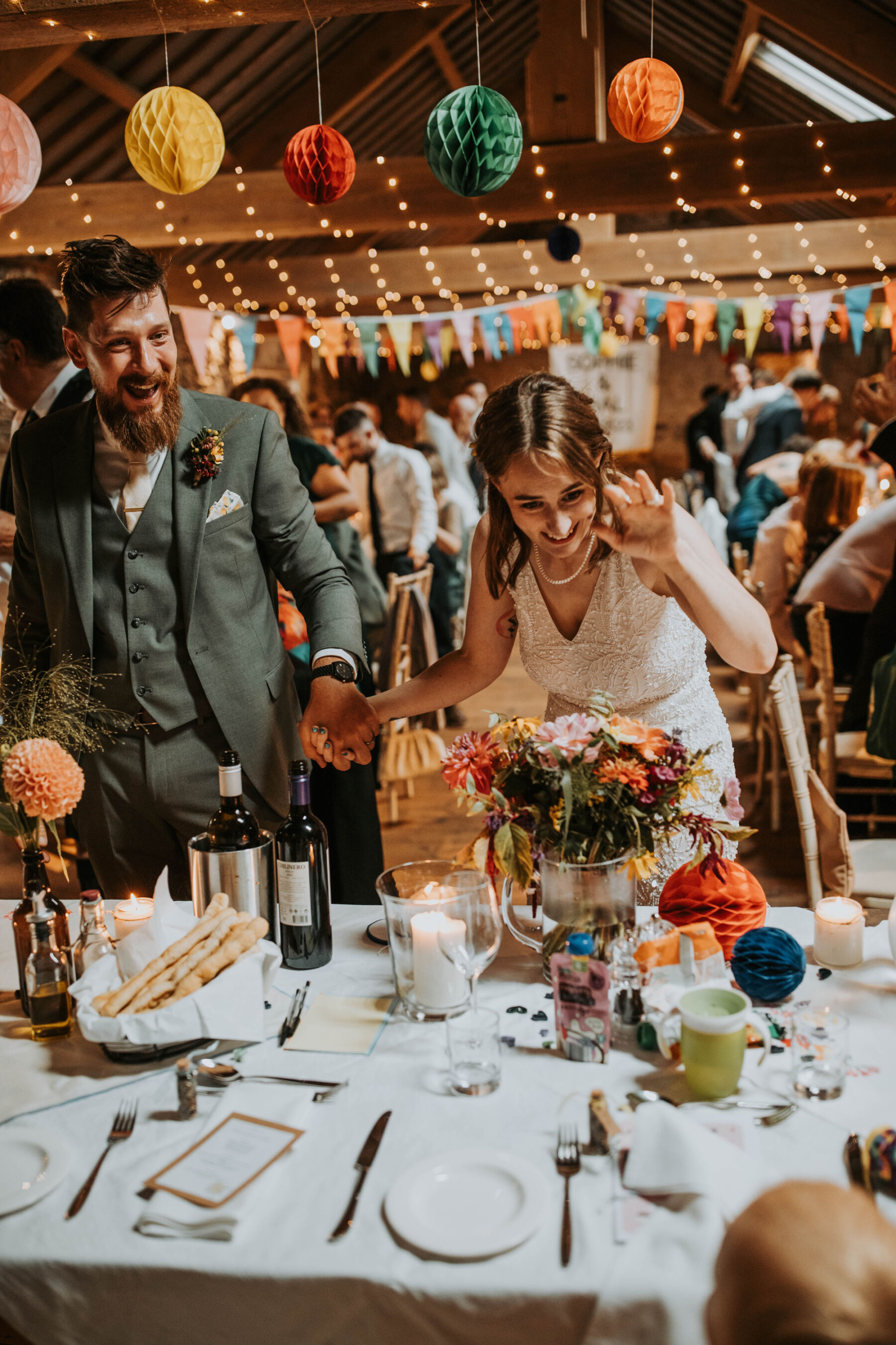 Sophie & Hal's Joyful, Colourful & Sustainable DIY Barn Wedding at Low Hall The Lakes 48 A delighted looking bride and groom arriving at their table inside the barn for their DIY wedding at Low Hall The Lakes.
