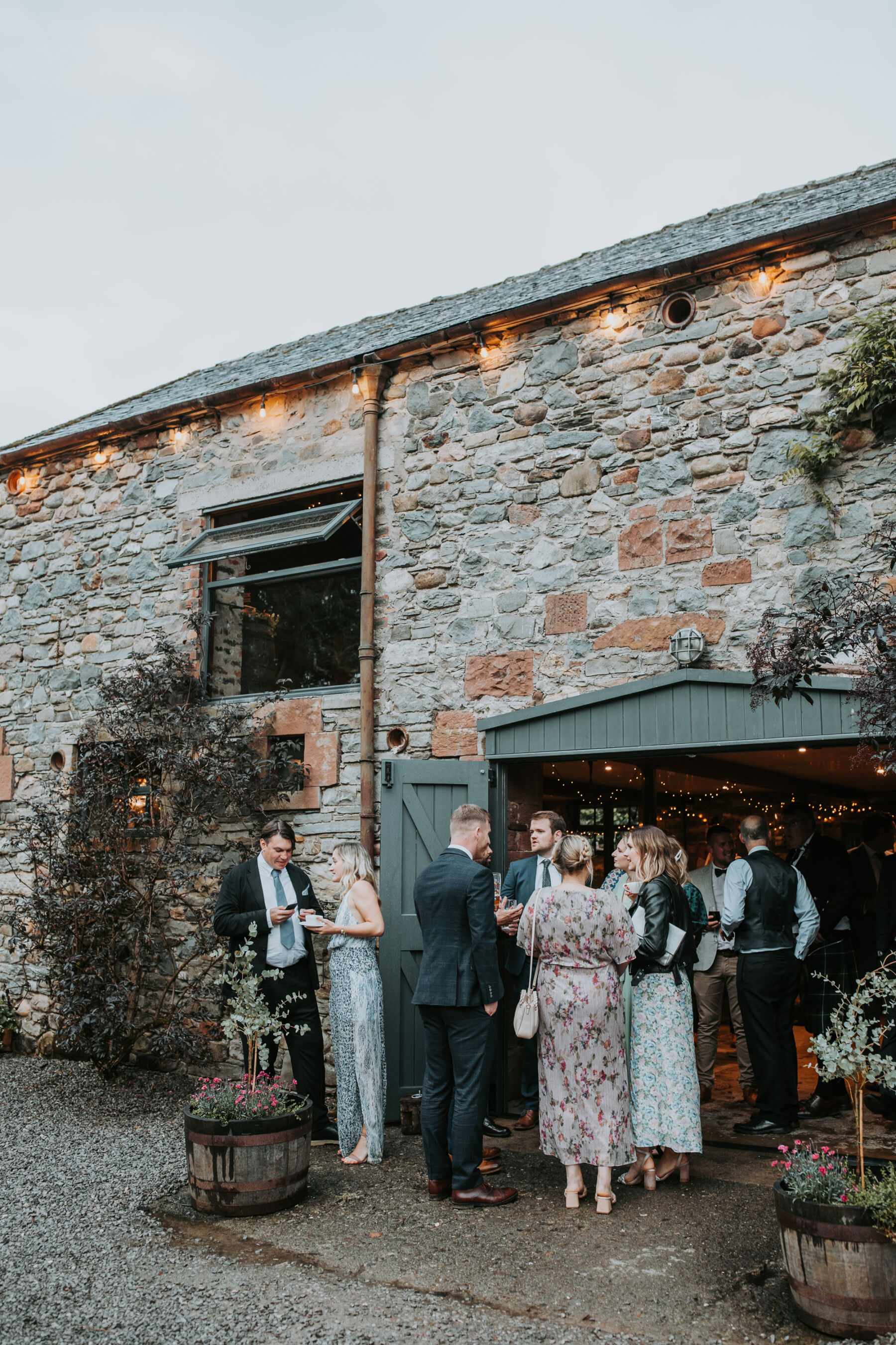 Sophie & Hal's Joyful, Colourful & Sustainable DIY Barn Wedding at Low Hall The Lakes 51 Wedding guests gathering outside Low Hall The Lakes wedding venue in The Lake District at dusk, with festoon lights lining the roof of the barn.