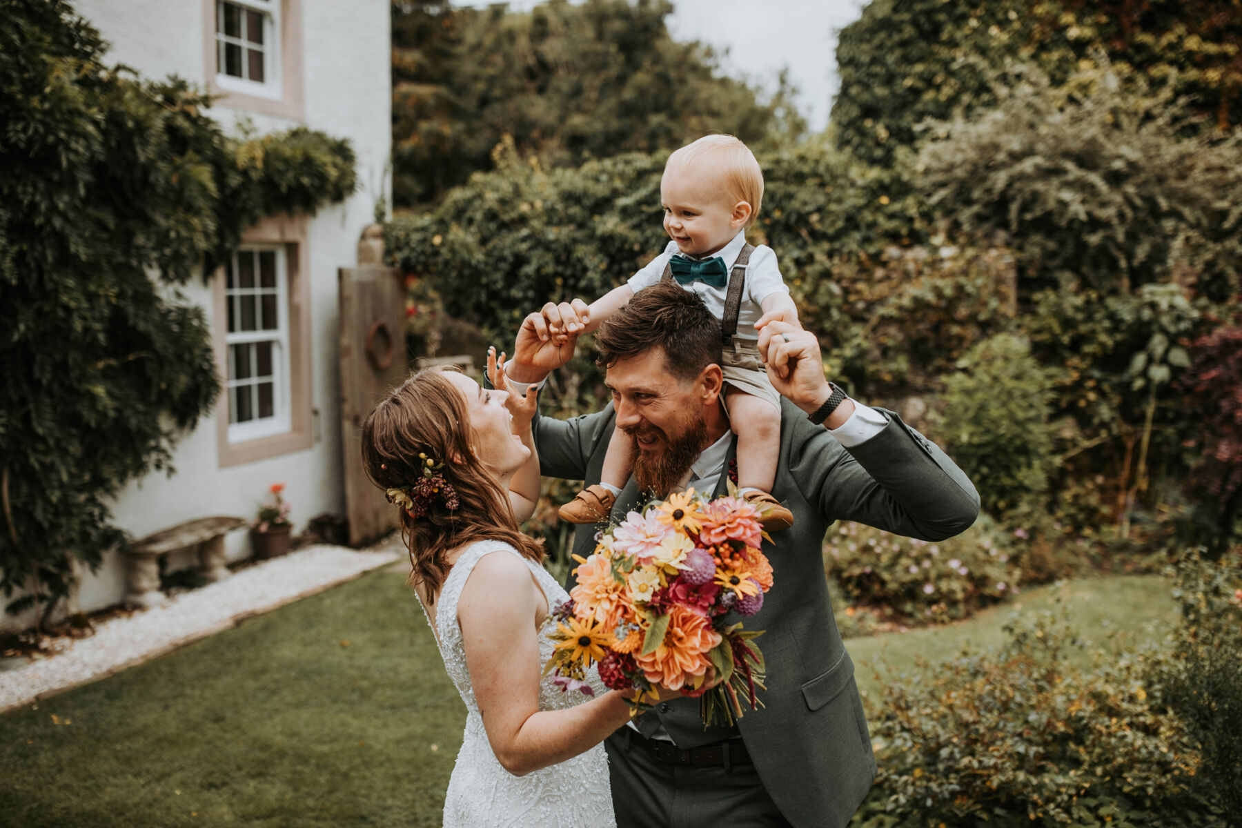 Sophie & Hal's Joyful, Colourful & Sustainable DIY Barn Wedding at Low Hall The Lakes 39 A joyful image of the bride, groom and their baby son sitting on the shoulders of the groom as the bride, with flowers in her hair and carrying a colourful summer bouquet, smiles and gesutres playfully to her son.