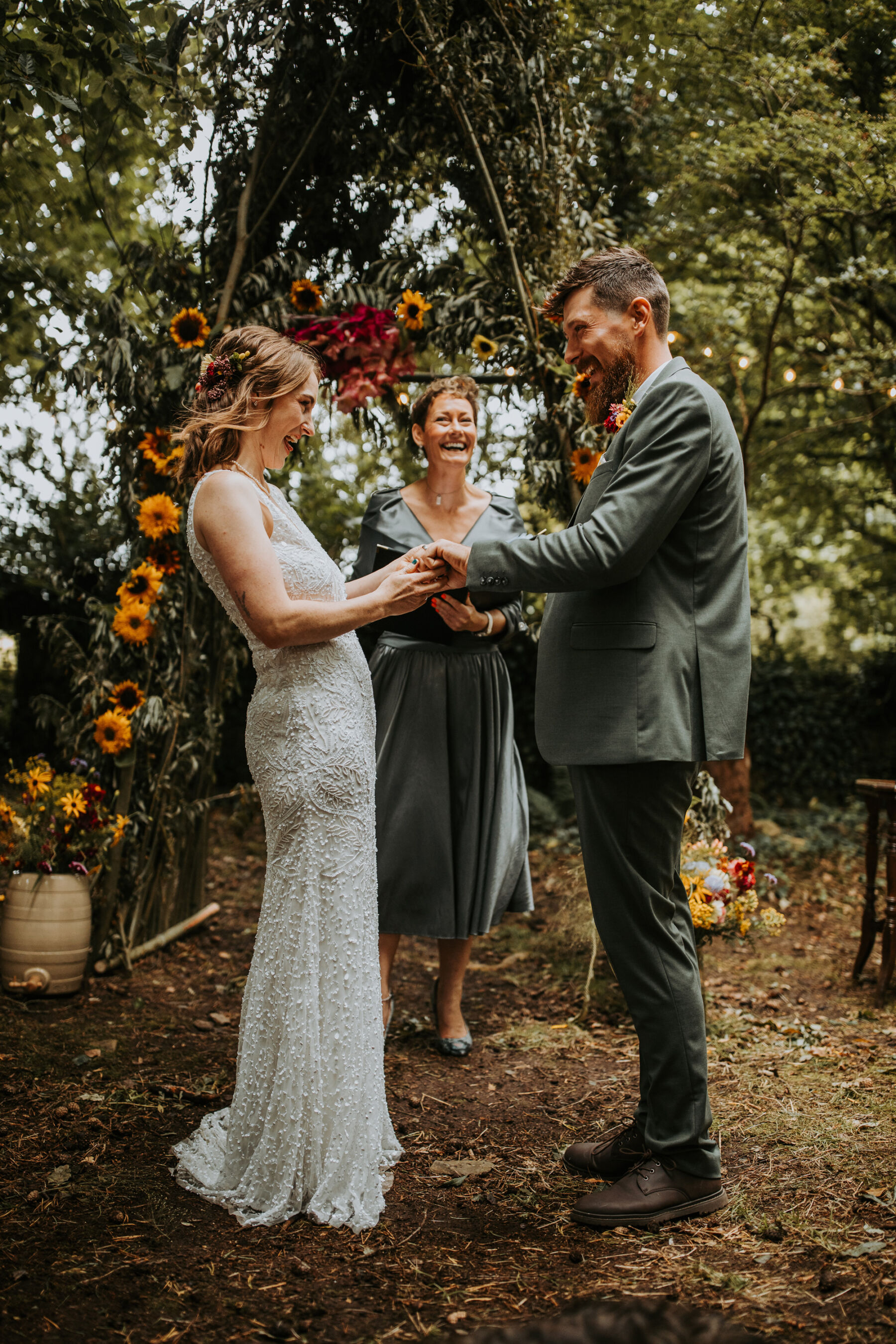 Sophie & Hal's Joyful, Colourful & Sustainable DIY Barn Wedding at Low Hall The Lakes 31 Kerrie of The Birds & The Bees Celebrant Services leading an outdoor wedding ceremony at Low Hall The Lakes in The Lake Distict. The Celebrant stands by an upside down V- shaped floral installation covered with sunflowers as the bride and groom hold hands standing in front of the Celebrant, and smile into each other's faces.