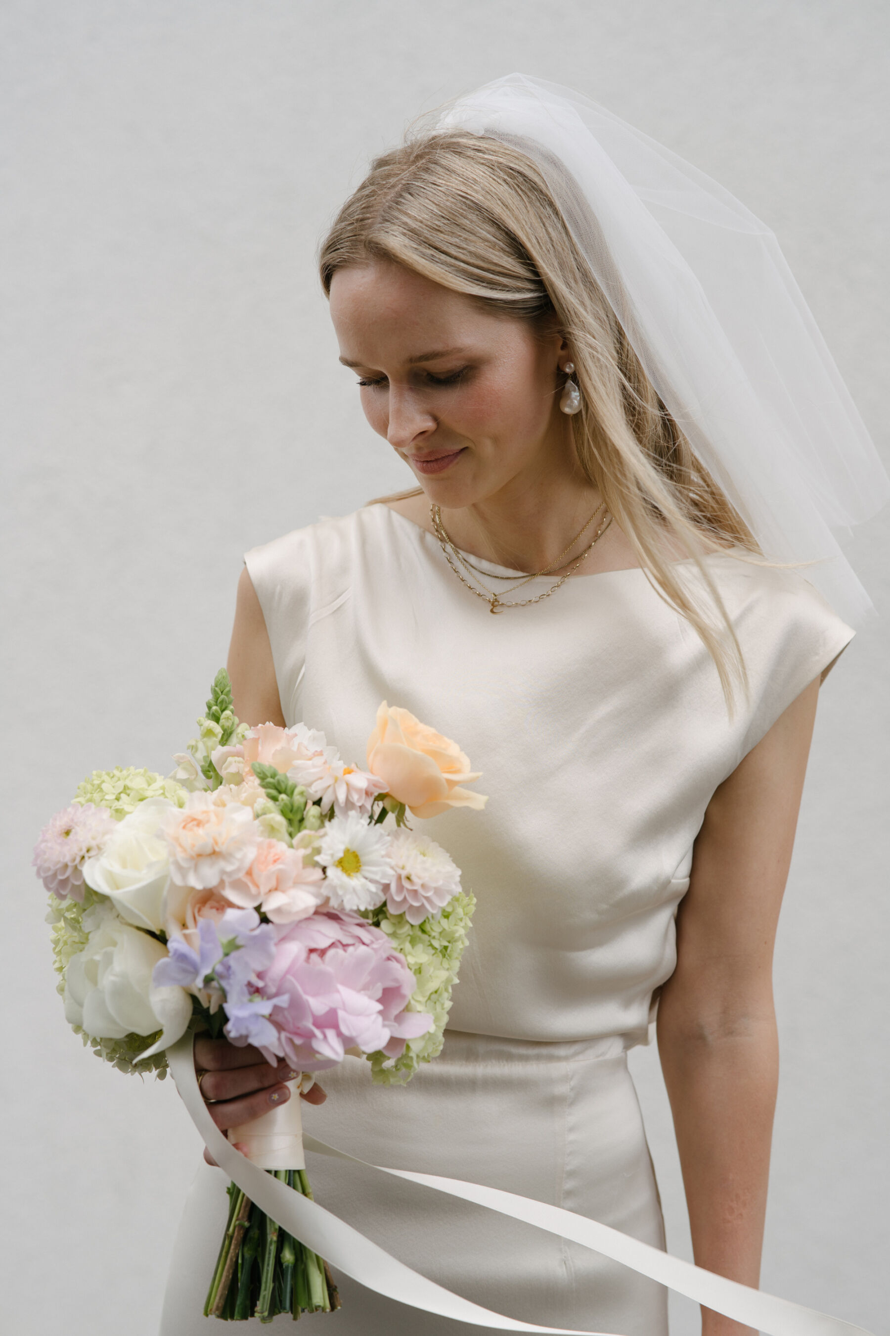 Relaxed, Romantic & Organic: Elena and Oliver's Cambridge Cottage Wedding at Kew Gardens 17 Modern bride wearing a Kamperett wedding dress and looking down on a beautiful pastel bridal bouquet with a silk ribbon.