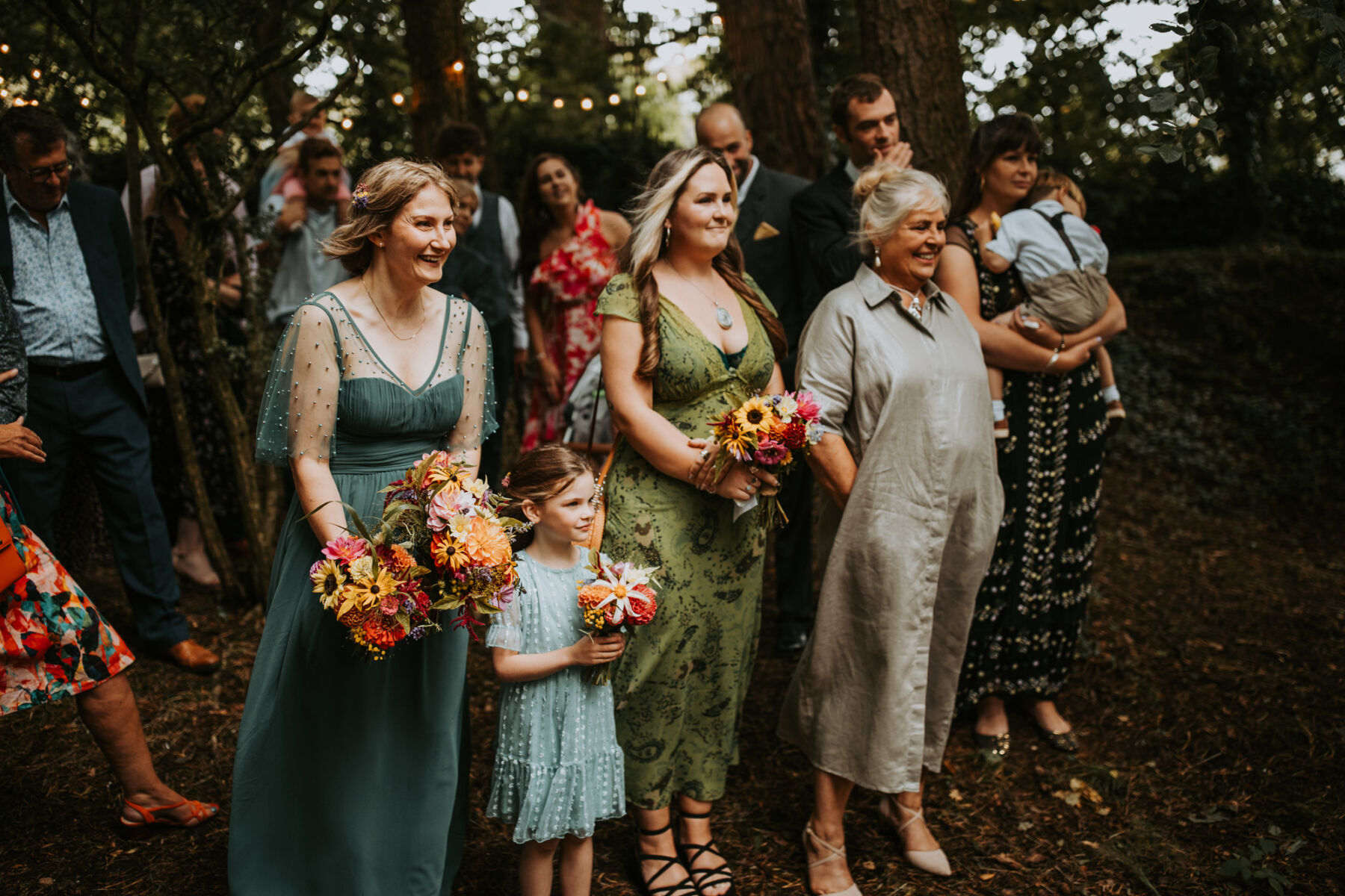 Sophie & Hal's Joyful, Colourful & Sustainable DIY Barn Wedding at Low Hall The Lakes 33 Photograph of the bridesmaids, flower girl and mother of the groom standing during an outdoor, woodland wedding ceremony at Low Hall The Lakes in The Lake District. The bridesmaids wear green.