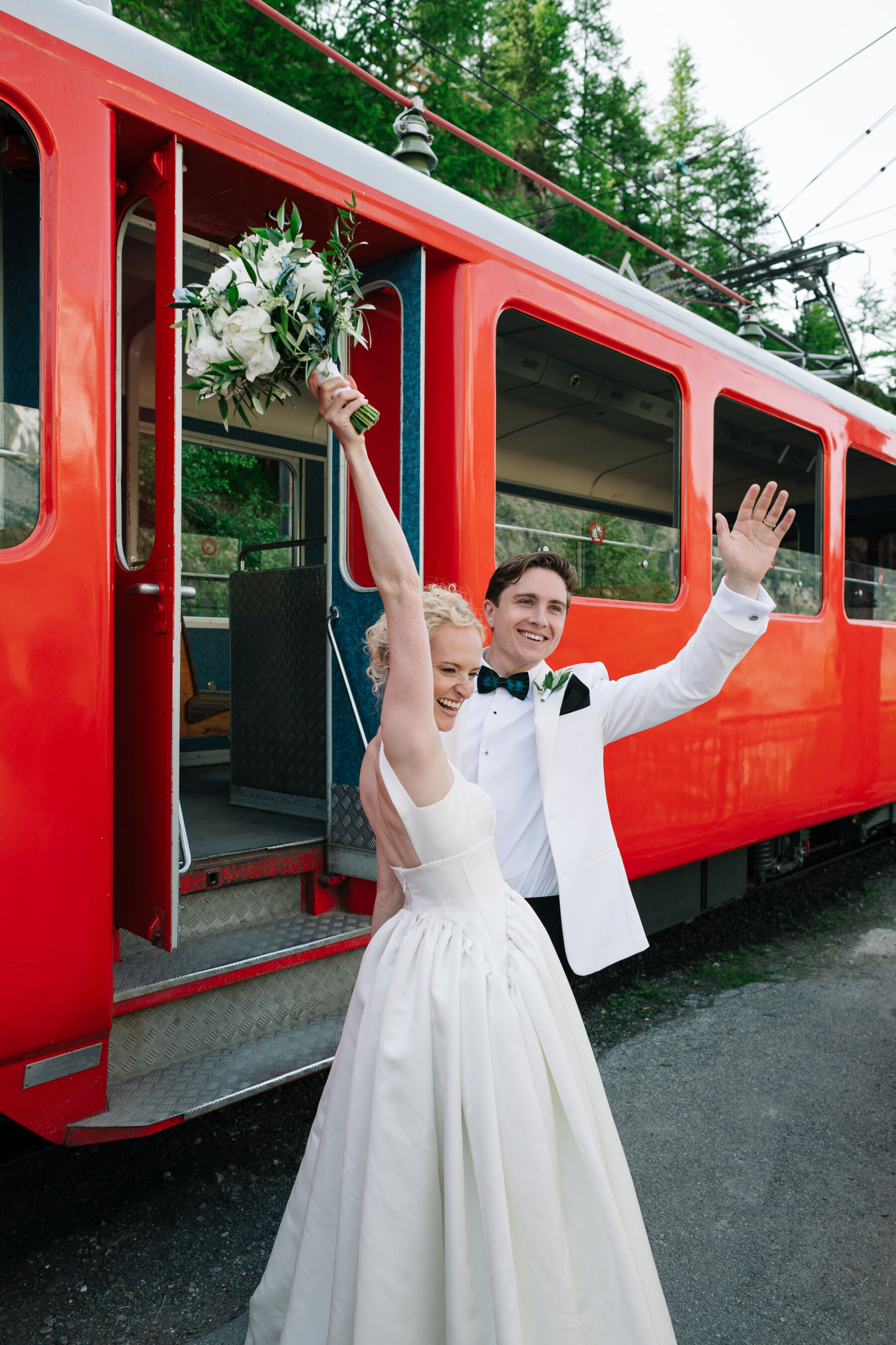 Georgia in Emilia Wickstead, For Her Classy & Elegant Black Tie Mountain Wedding in the Chamonix 4 Bride in an Emilia Wickstead wedding dress, raising her bouquet of white flowers into the air, her groom in black tie and white tuxed, they stand next to a red colour ski train in the Chamonix mountains.