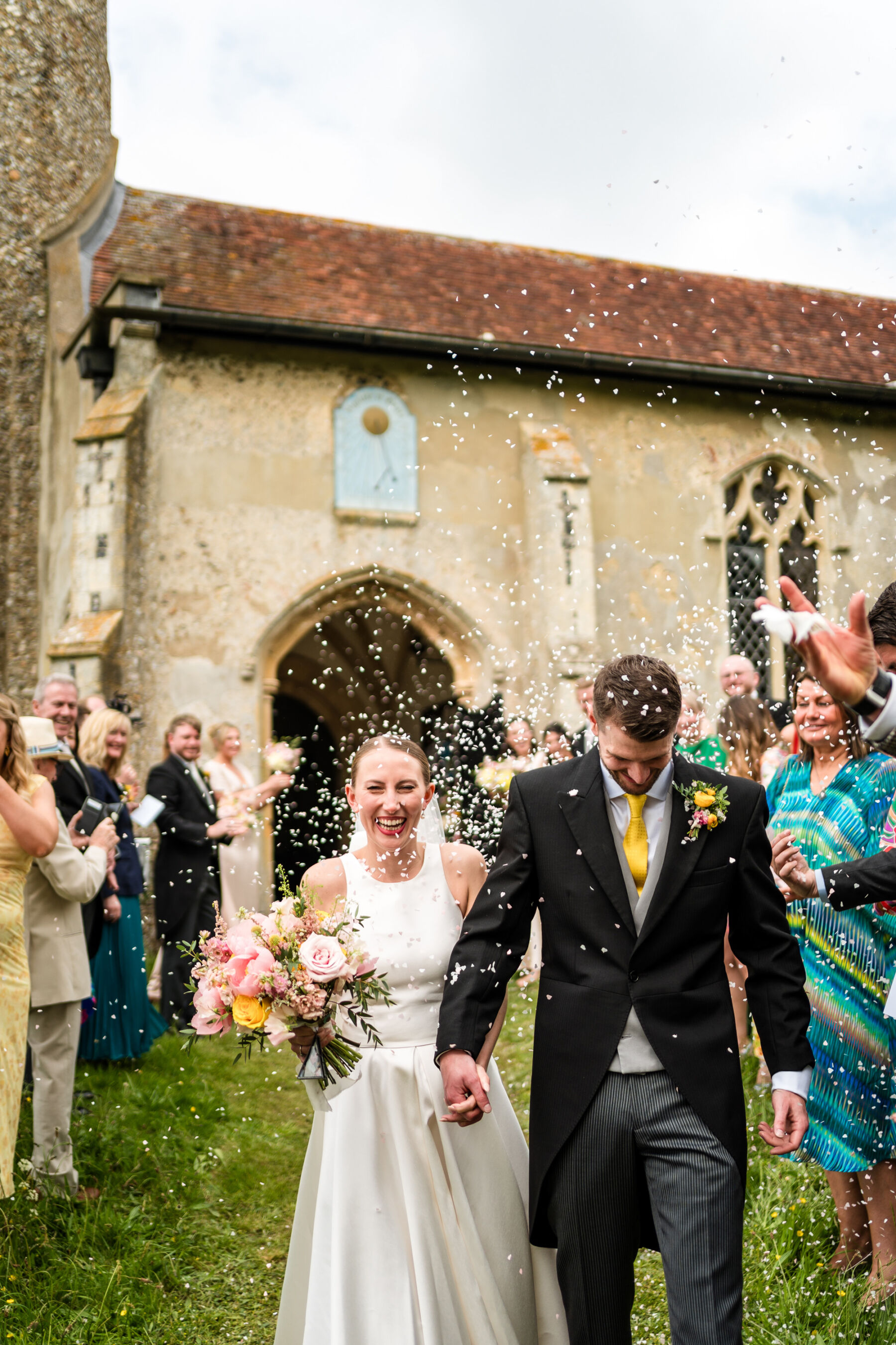 Alice Wore Oak by Halfpenny London For Her Charming Spring Marquee Wedding in Suffolk 22 Confetti moment outside the church. Bride wears the Oak by Halfpenny London wedding dress, and carries a colourful bouquet of wedding flowers. The groom wears a traditional morning suit with a yellow tie. Yellow flowers feature in the brides bouquet and groom's button hole.