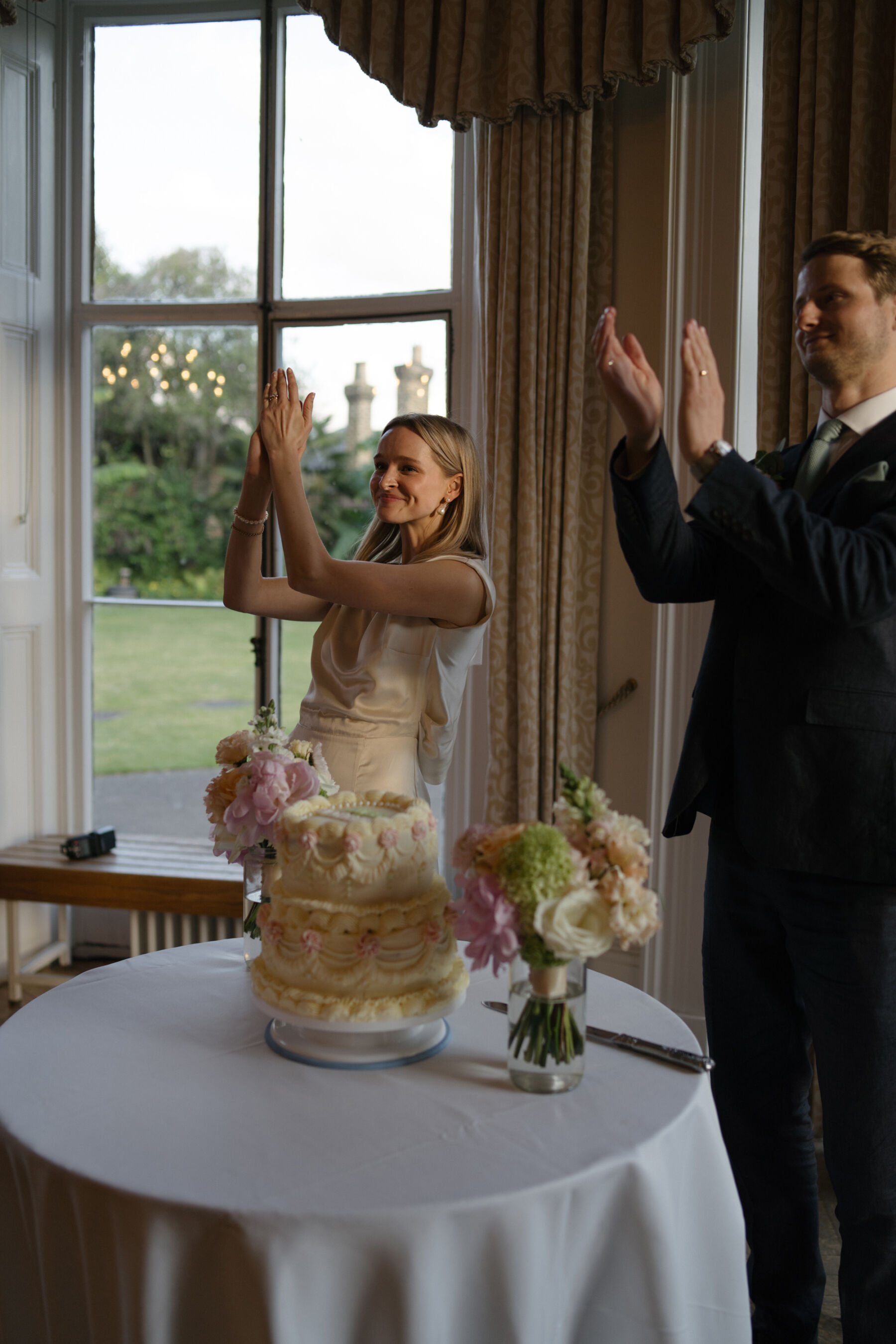 Relaxed, Romantic & Organic: Elena and Oliver's Cambridge Cottage Wedding at Kew Gardens 33 Bride and groom raising their hands to clap as they stand near to their 3 tier wedding cake with frilly icing.