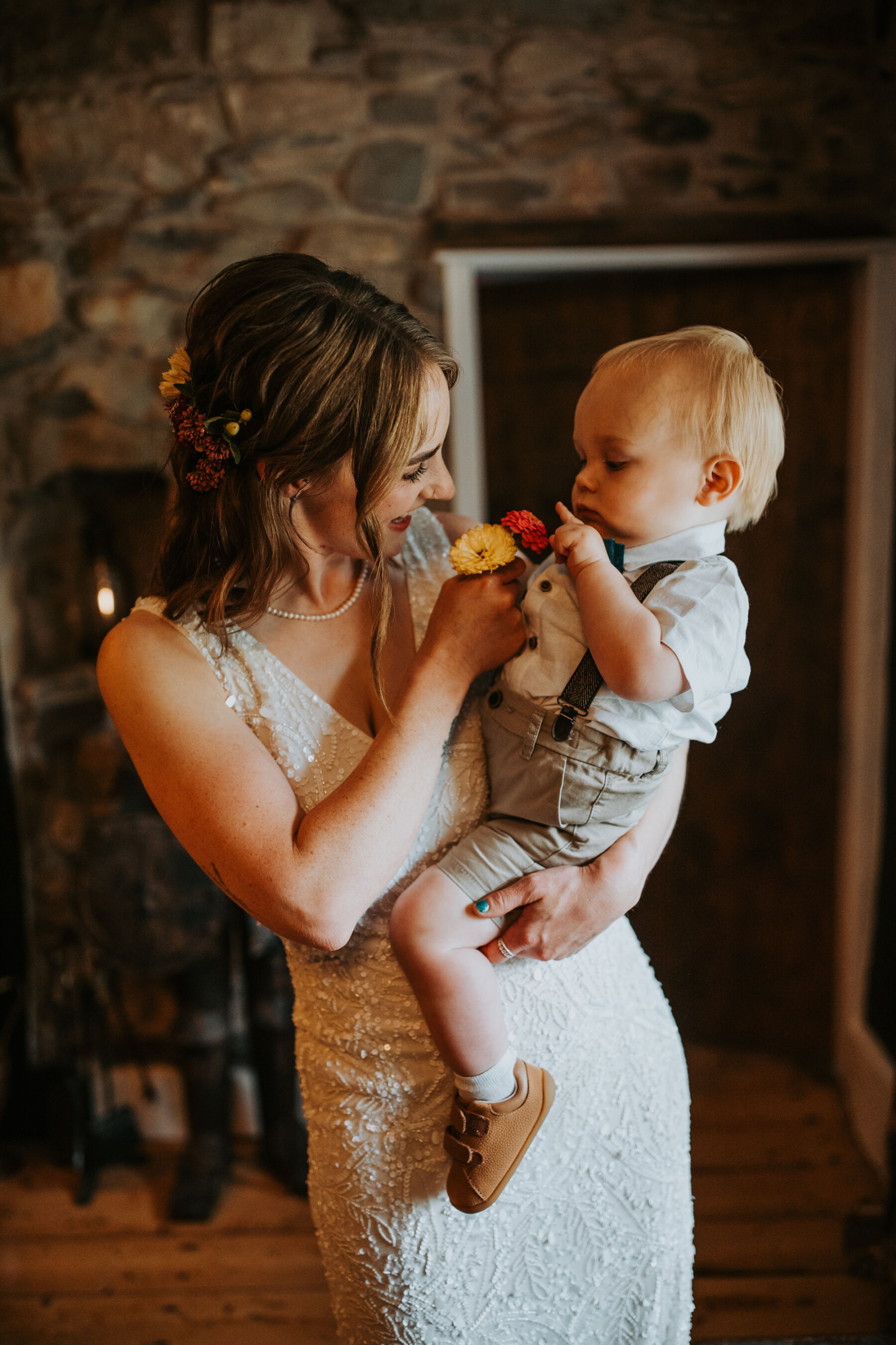Sophie & Hal's Joyful, Colourful & Sustainable DIY Barn Wedding at Low Hall The Lakes 28 A bride with loose, shoulder length hair and flowers in her hair, wearing a sequin ASOS wedding dress, carrying her baby son on her waist who is wearing shorts, braces and a green velvet bow tie.