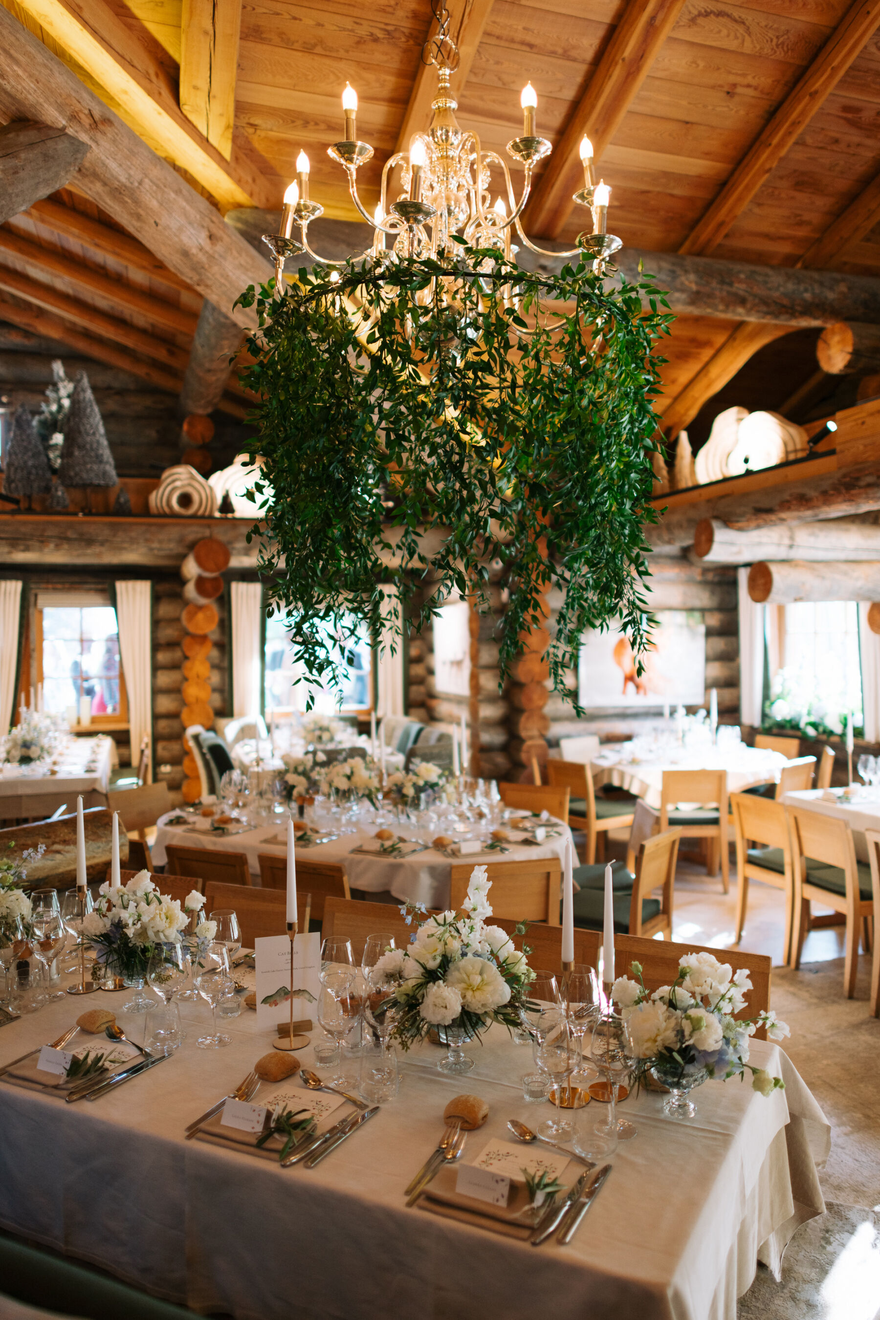 Georgia in Emilia Wickstead, For Her Classy & Elegant Black Tie Mountain Wedding in the Chamonix 33 Greenery hanging from a candle chandelier that hangs above a round table in a wedding reception. Below are bowls of white flowers and tall white taper candles in slim, modern, gold candle holders.