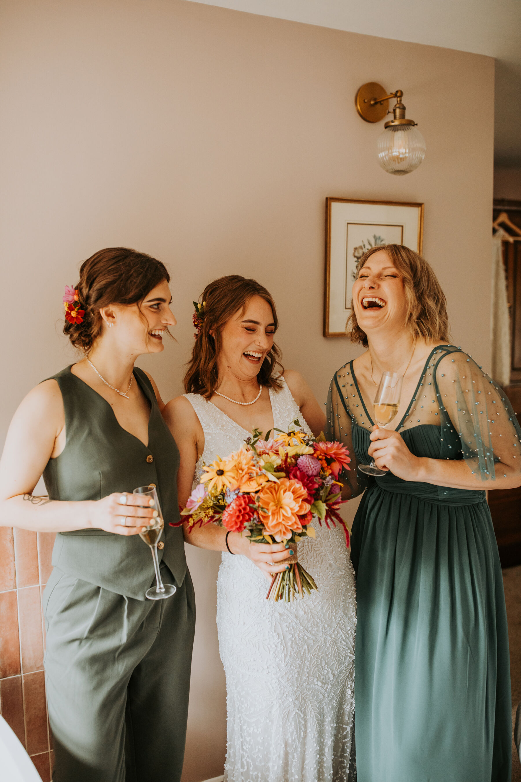 Sophie & Hal's Joyful, Colourful & Sustainable DIY Barn Wedding at Low Hall The Lakes 24 Bride standing in the middle of two bridesmaids in green. The bride leans back with laughter as she holds a bright and colourful wedding bouquet.
