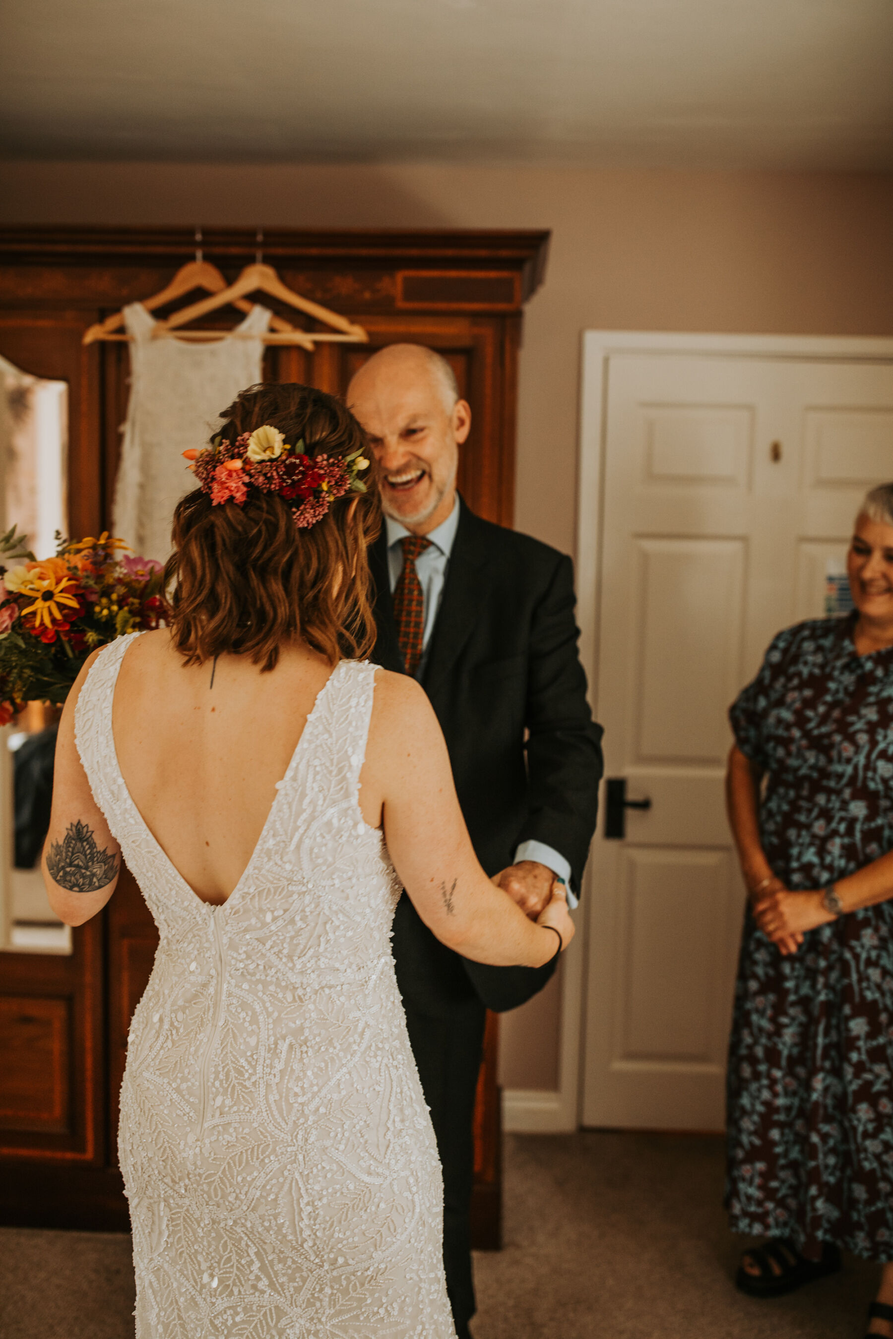 Sophie & Hal's Joyful, Colourful & Sustainable DIY Barn Wedding at Low Hall The Lakes 16 The bride's father seeing his daughter for the first time on her wedding. He looks happy and delighted. The bride has her back to the camera and you can see colourful flowers in her loose, shoulder length hair.
