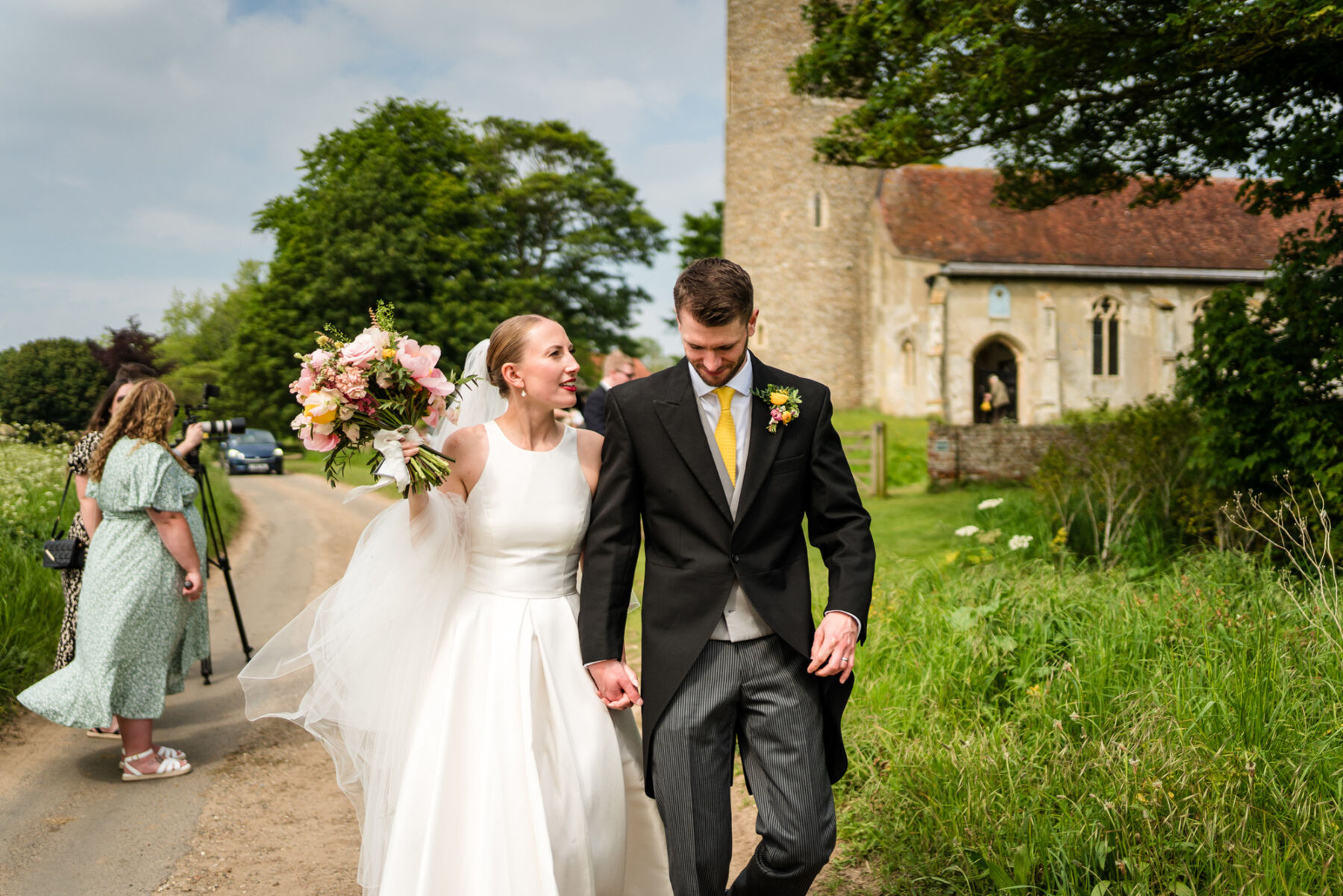 Alice Wore Oak by Halfpenny London For Her Charming Spring Marquee Wedding in Suffolk 27 Bride and groom holding hands as they walk away from the church. The bride carries a colourful bouquet and wears the Oak dress by Halfpenny London. The groom wears traditional morning suit and yellow tie.