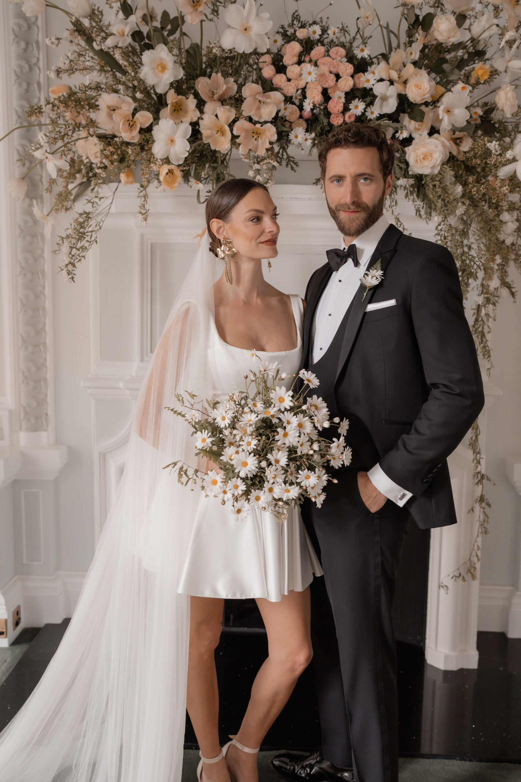 Bride wearing a short and playful wedding dress by Halfpenny London, and carrying a wedding bouquet of Oxeye Daisies. Behind her and her groom in black tie is a fireplace adored 
 with peach flowers.