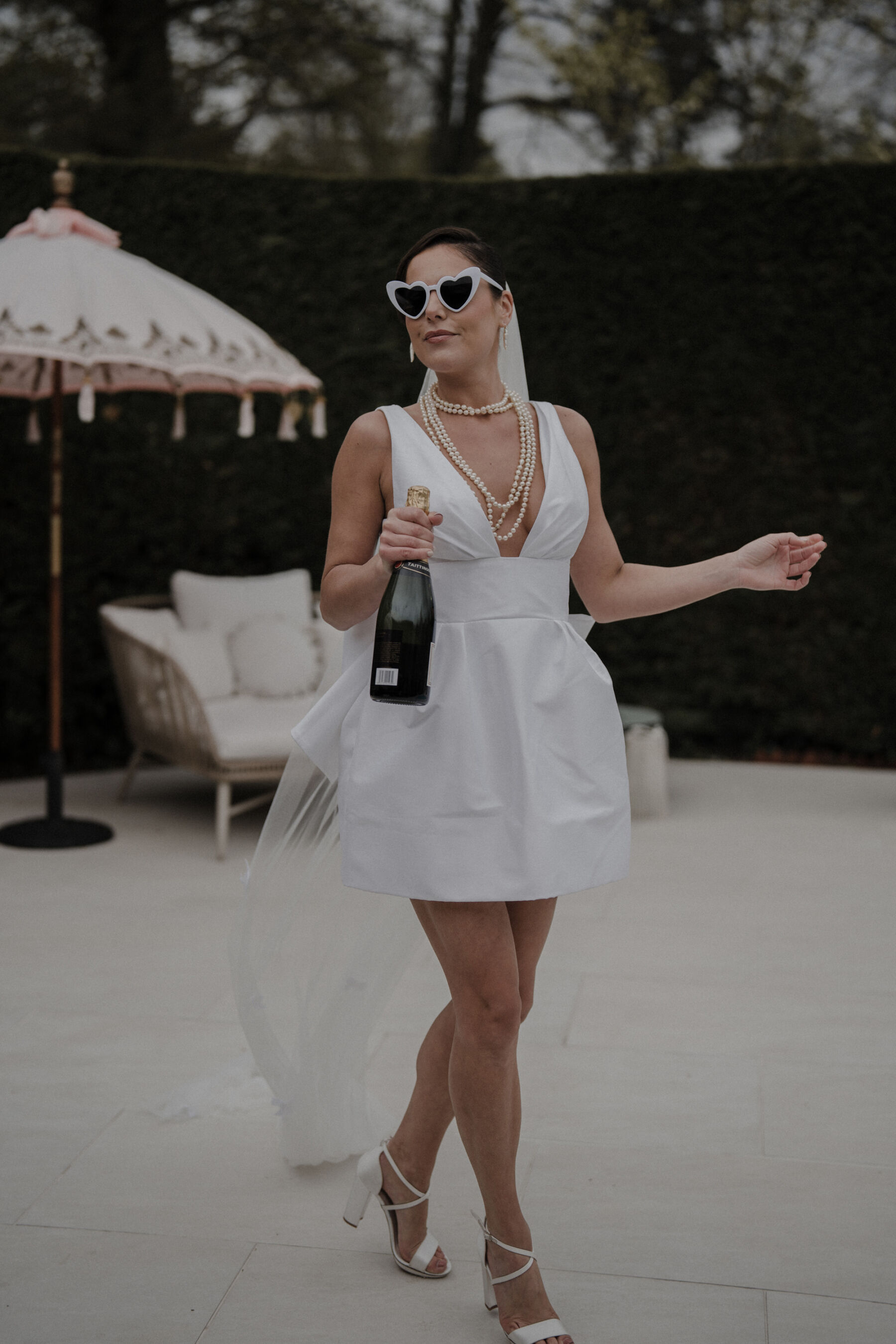 Bride clutching a bottle of champagne and wearing pearls, a short wedding dress, block heels and heart shaped sunglasses. Chic and sassy bridal look.