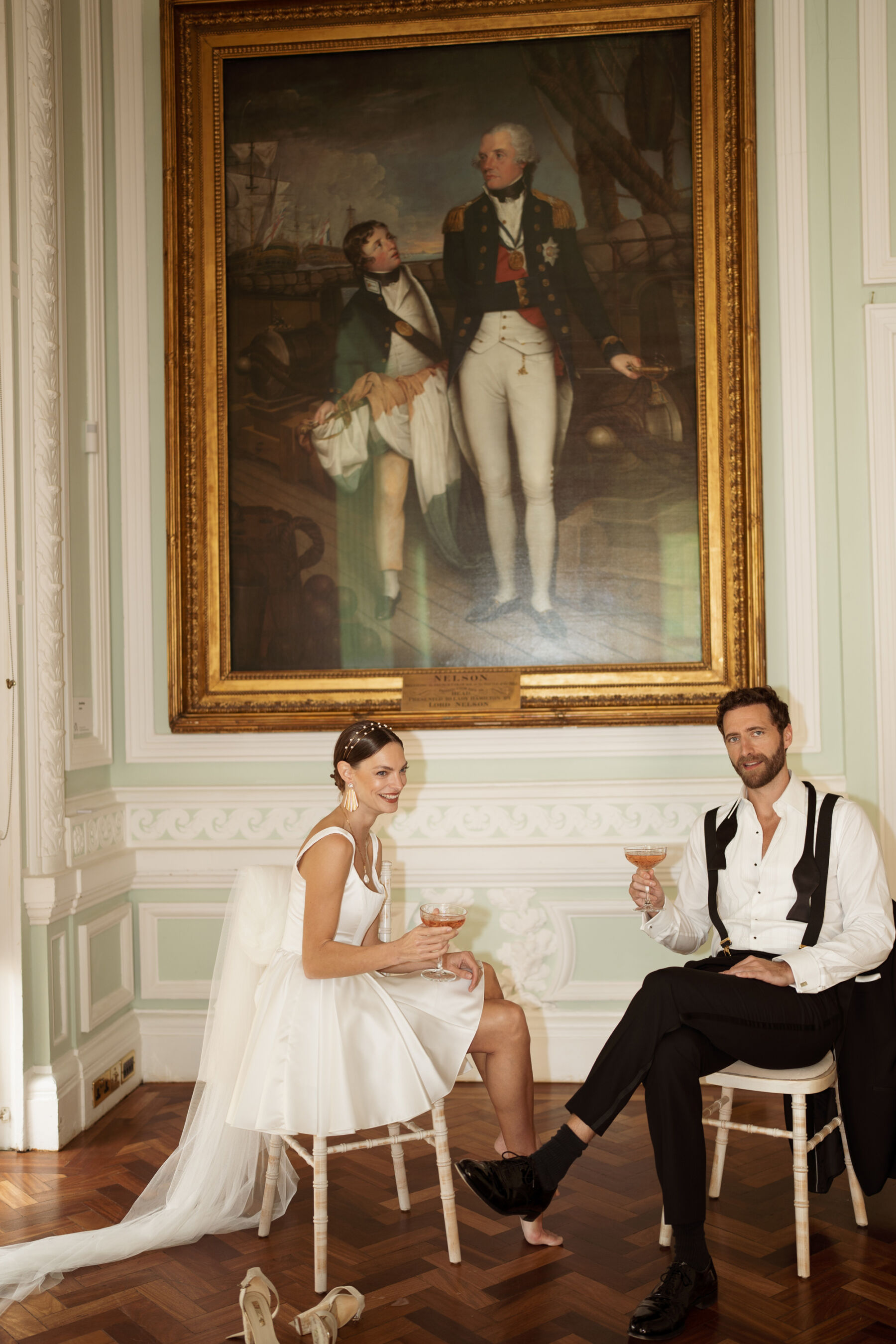 Bride and groom seated in relaxed poses beneath a painting on the wall at 10-11 Carlton Terrace. The groom has loosened his bowtie and unbuttoned his shirt and the bride has taken off her shoes. They are in playful mood.