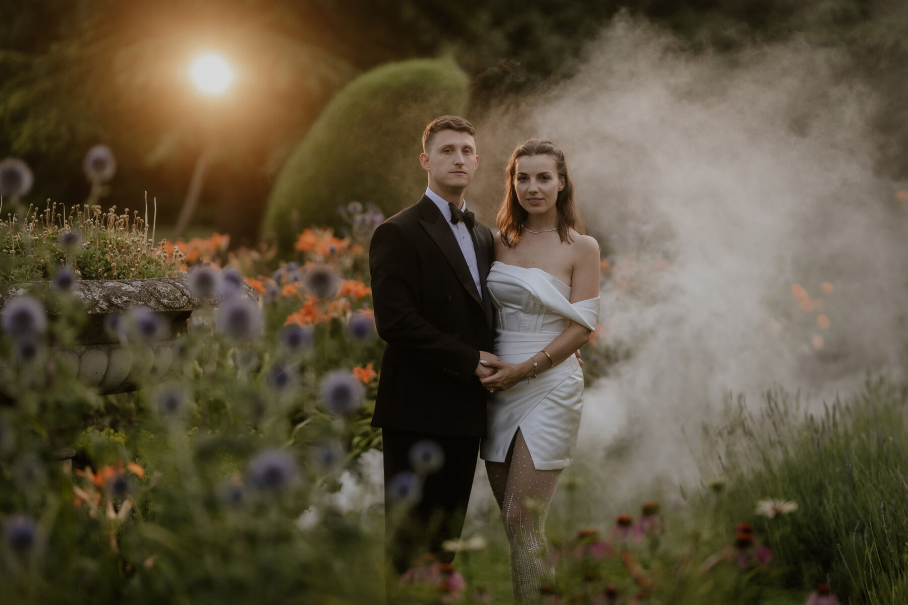 An Investment Banker & Her Prestwold Hall Wedding, with a Milla Nova Gown & Berta Legging Boots 42 Bride in a mini dress and Berta legging Boots. Groom in black tie and pale blue braces. The couple stand amidst smoke in the grounds of Prestwold Hall.