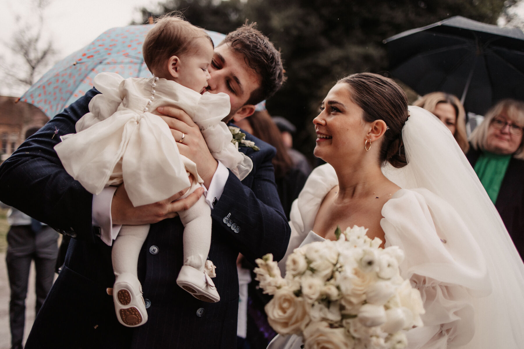 Maddy & Nik’s Cosy and Intimate Winter Wedding with a Custom Jesus Peiro Gown 17 Groom holding is sweet baby daughter who wears a little silk ivory dress, white tights and white shoes with bows. Her mother looks up at her lovingly.