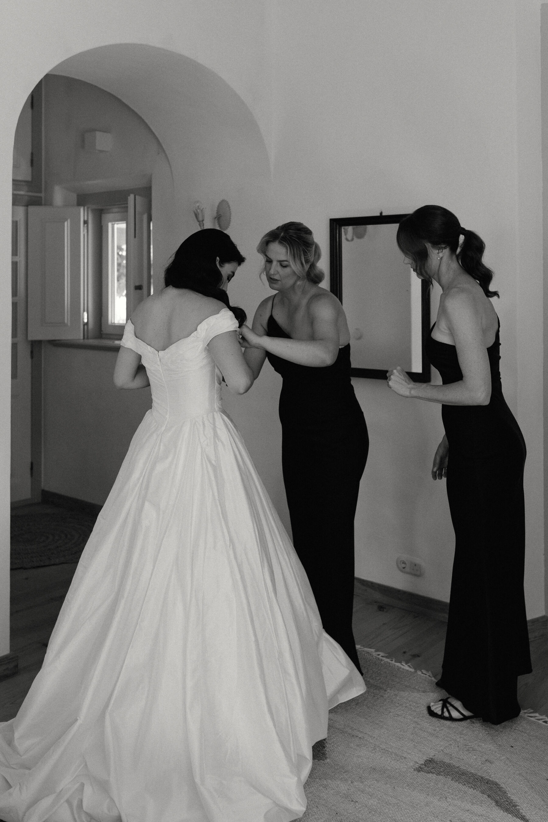 Olivia & Tim's Classy & Elegant Vineyard Wedding in Portugal 19 Two bridesmaids in black dresses assisting the bride into her wedding dress.