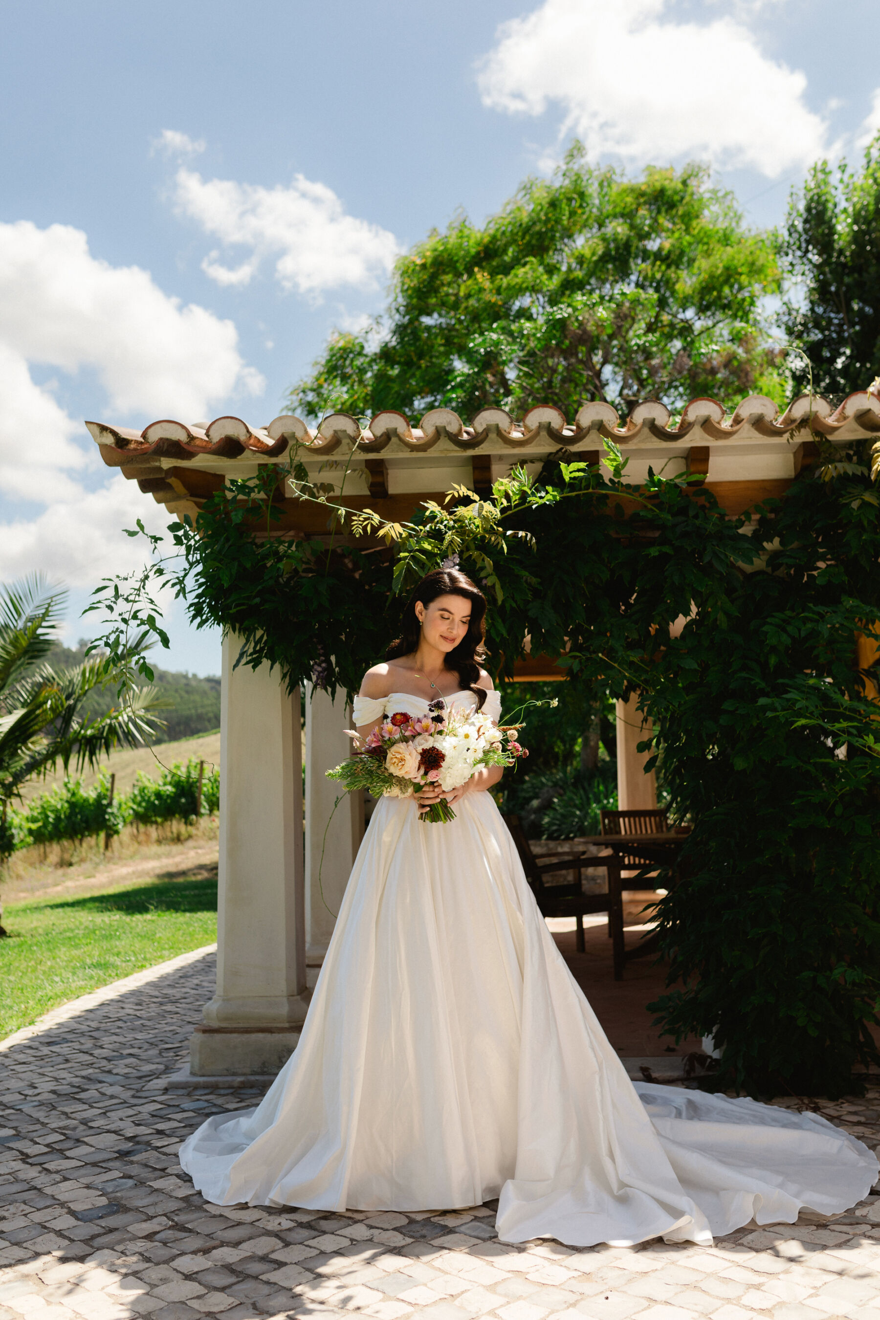 Olivia & Tim's Classy & Elegant Vineyard Wedding in Portugal 21 Bride with long dark wavy hair and an elegant off the shoulder dress with a large train, carrying a colourful and elegant summer wedding bouqet.
