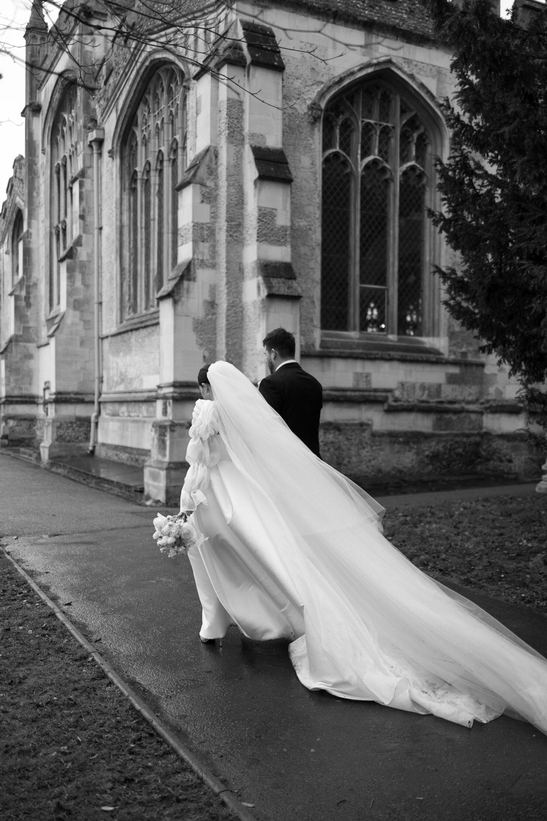 Maddy & Nik’s Cosy and Intimate Winter Wedding with a Custom Jesus Peiro Gown 22 Bride and groom holding hands as they stroll by the church. The groom wears a navy suit and pale blue tie. The bride wears a Jesus Peiro ballgown style wedding dress with puff sleeved cape and veil. She wears Prada pointed stilettos.
