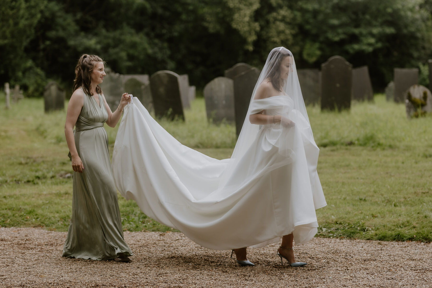 An Investment Banker & Her Prestwold Hall Wedding, with a Milla Nova Gown & Berta Legging Boots 18 Bridesmaid in a sage green dress lifting the dress of the bride as they walk to the church. The bride wears Milla Nova.