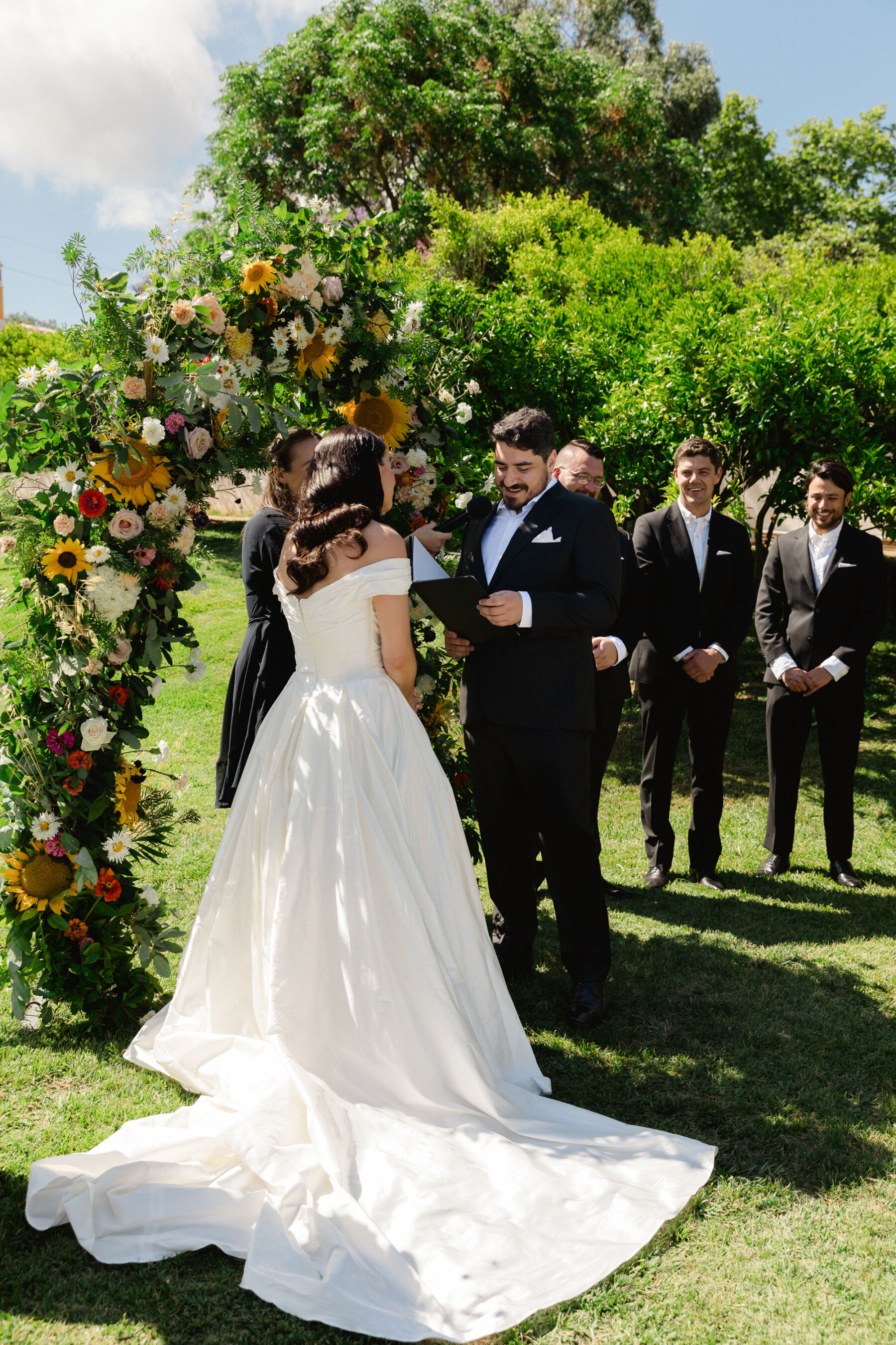 Olivia & Tim's Classy & Elegant Vineyard Wedding in Portugal 25 Bride and groom standing by a large floral arch with sunflowers and Dahlia, for their outdoor wedding in Portugal.
