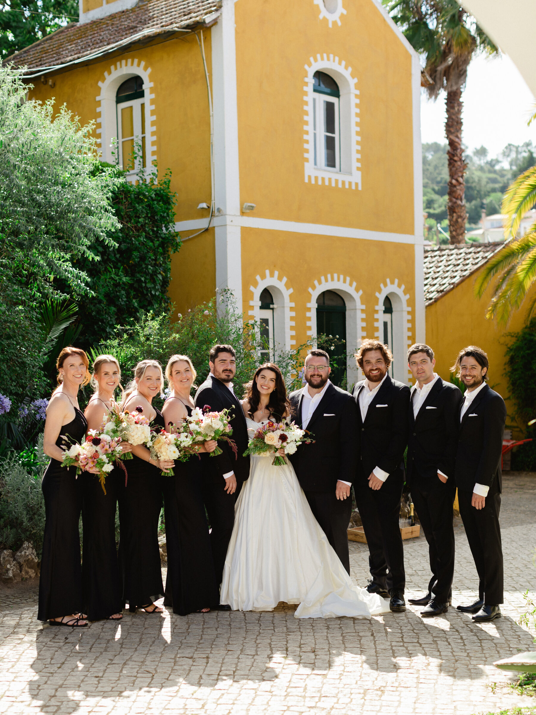 Olivia & Tim's Classy & Elegant Vineyard Wedding in Portugal 29 Classy and elegant destination wedding. Bride and groom standing by a yellow building in Portugal with their bridesmaids and groomsmen dressed all in black.