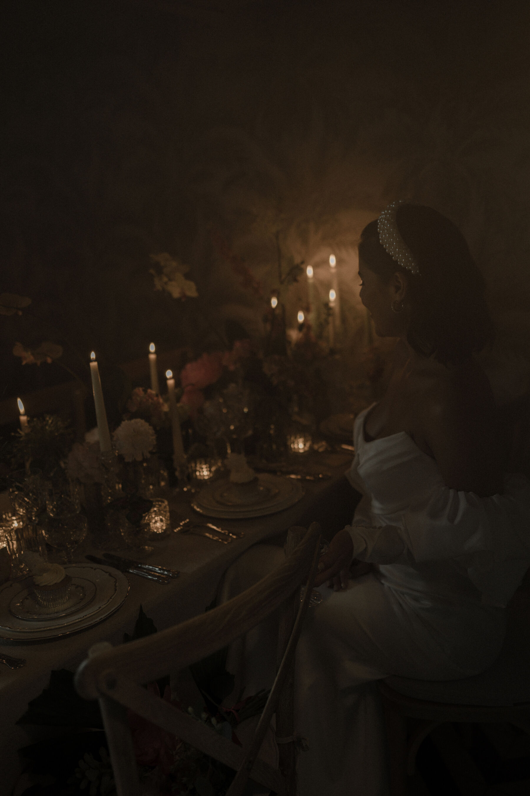 Dark, moody and romantic image of a bride sat at a candlelit table with peonies.