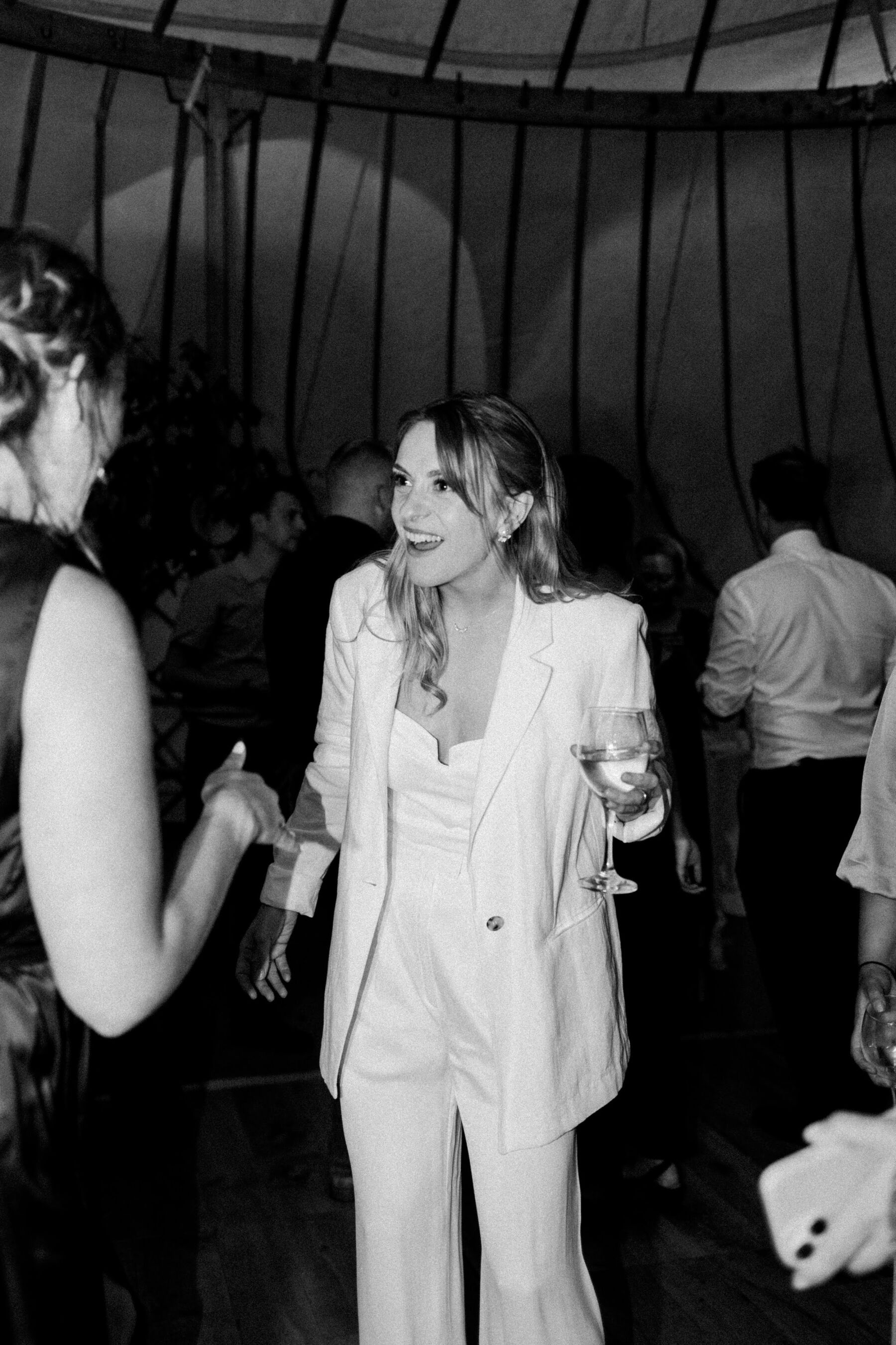 A Summer Yurt Wedding at The Bride's Parent's Home in Northumberland 45 Bride in a white jacket and wide legged trousers clutching a glass of champagne.