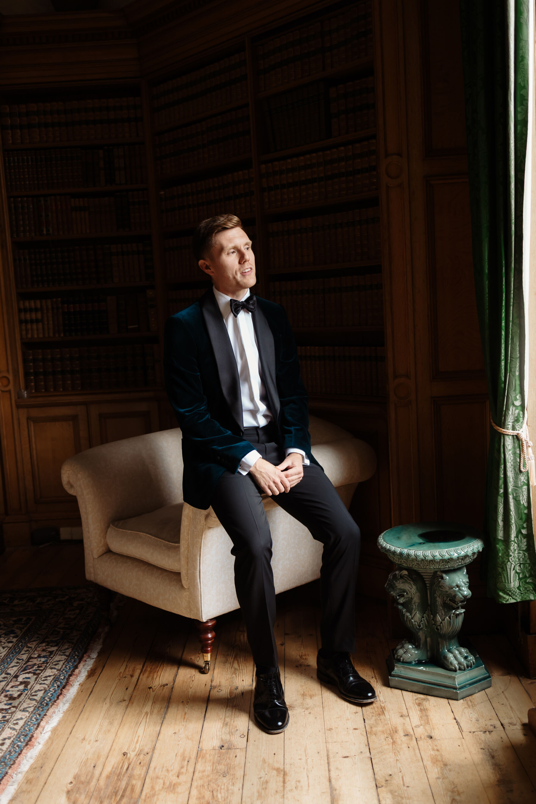 Emily in Rosa Clara for her Romantic & Elegant July Wedding at Cluny Castle 14 Groom sat on the edge of a chair at Cluny Castle wedding venue in Aberdeenshire. He wears a dark blue tuxedo and bow tie.