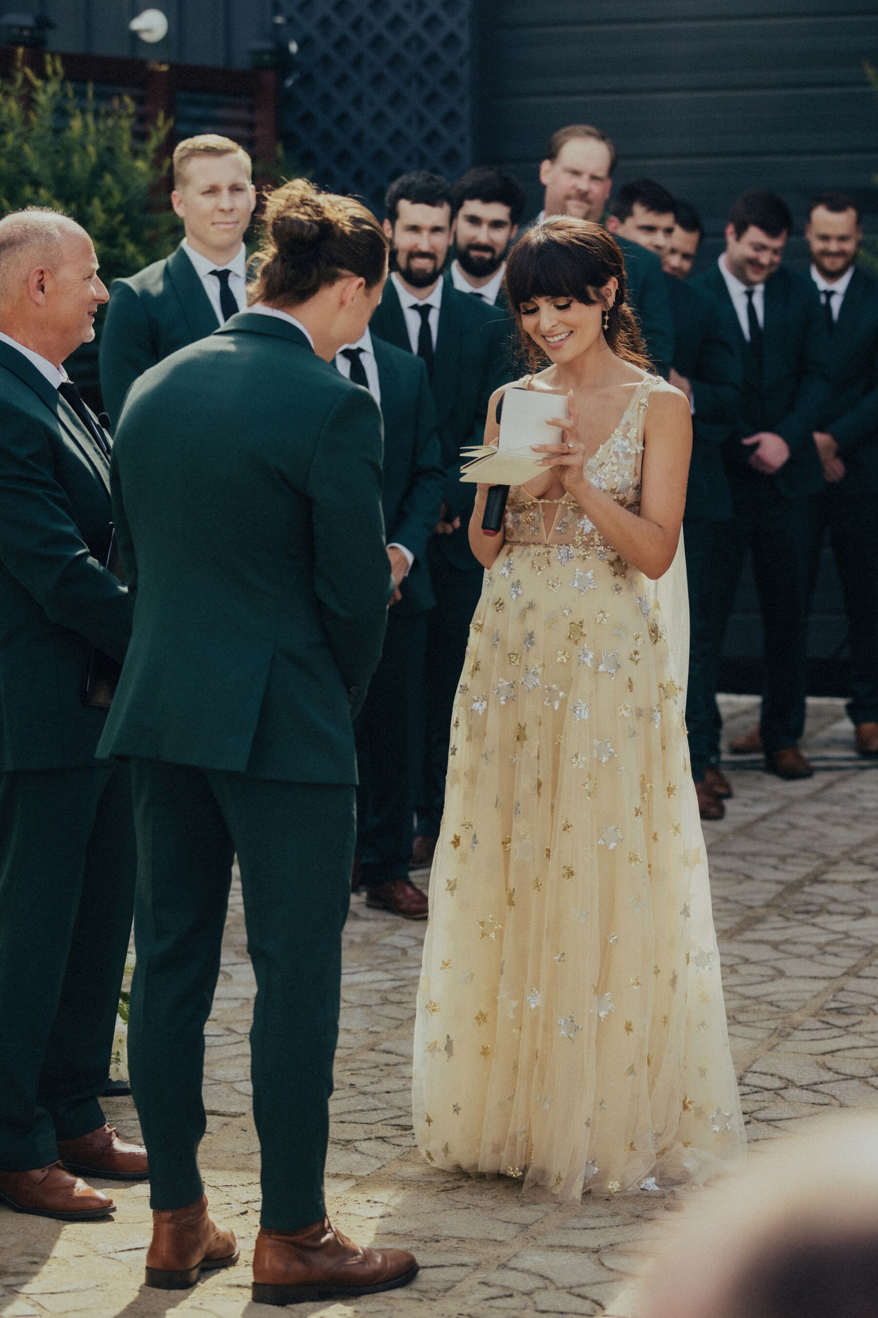 Bride and groom exchanging vows during their outdoor wedding. The groom wears a dark green suit, the bride wears the Orion dress, a celestial inspired wedding dress by Watters. The bride is smiling as she looks down at her written vows held in her hand.