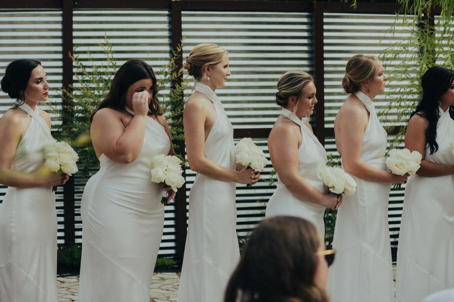 Emotional bridesmaids standing during the wedding ceremony and crying happy tears and wiping tears from their eyes. They wear halterneck white dresses.