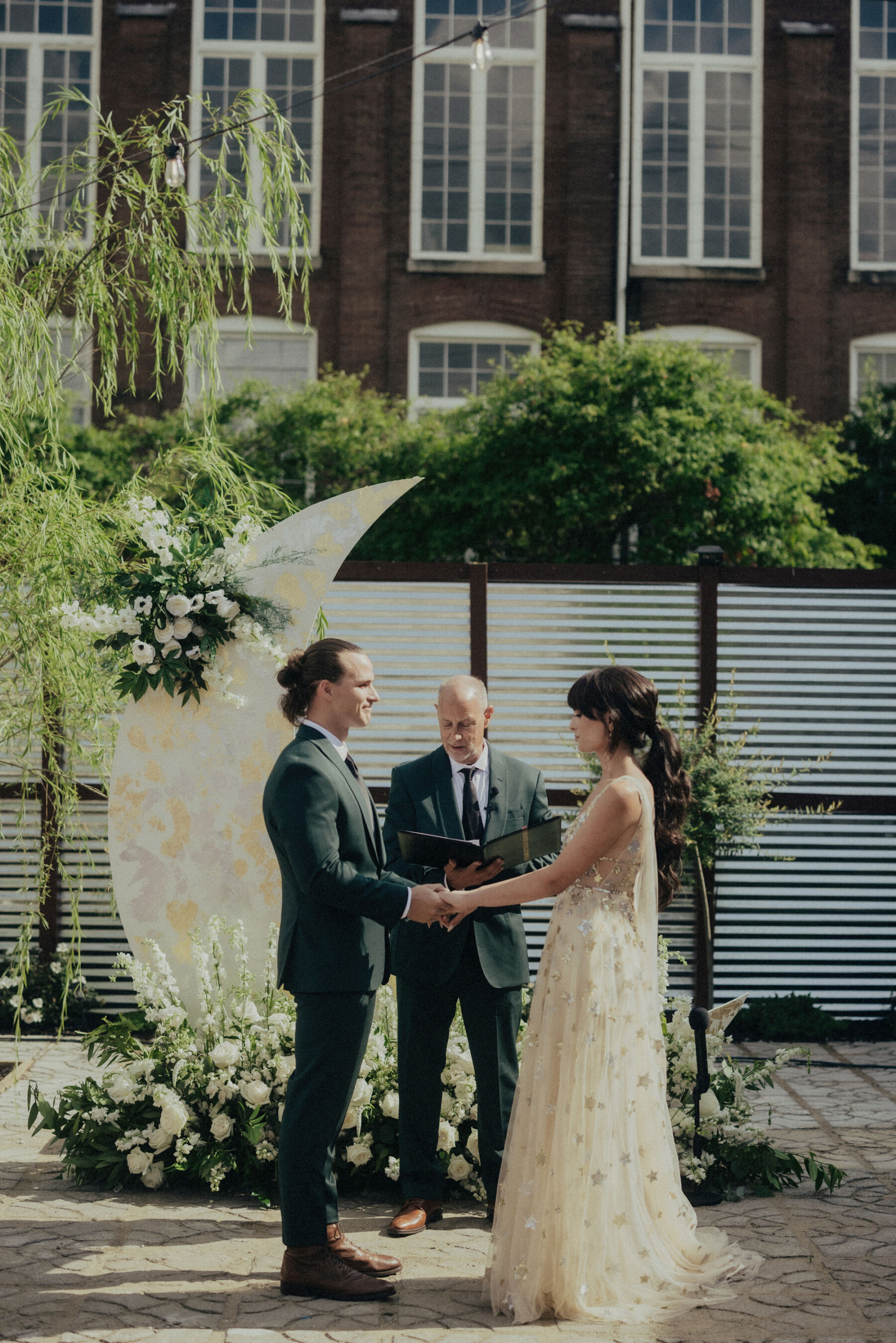 Bride and groom holding hands as they get married. Behind them is a crescent moon prop decorated with flowers. The groom wears a dark green suit, the bride wears the Orion dress, a celestial inspired wedding dress by Watters.