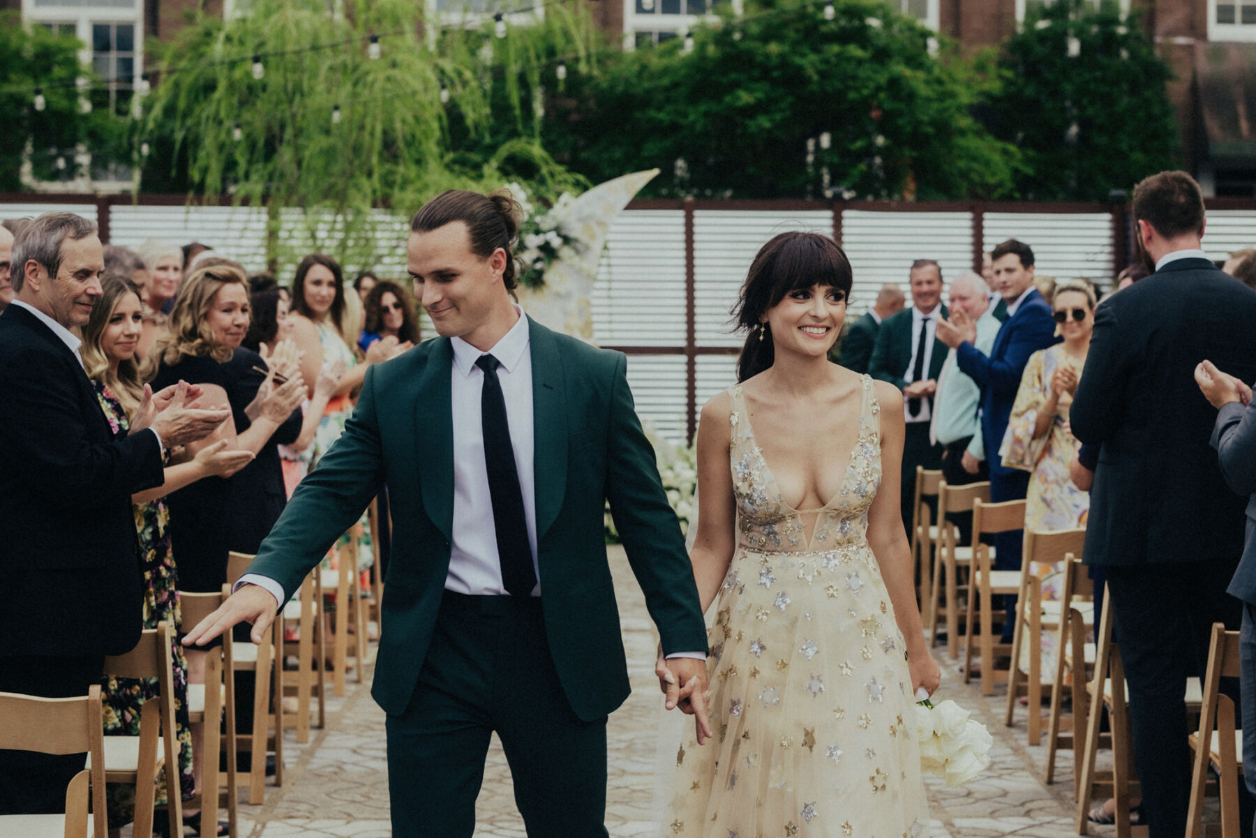Groom in dark green suit holding hands with his bride in a celestial wedding dress as they walk back down the aisle.