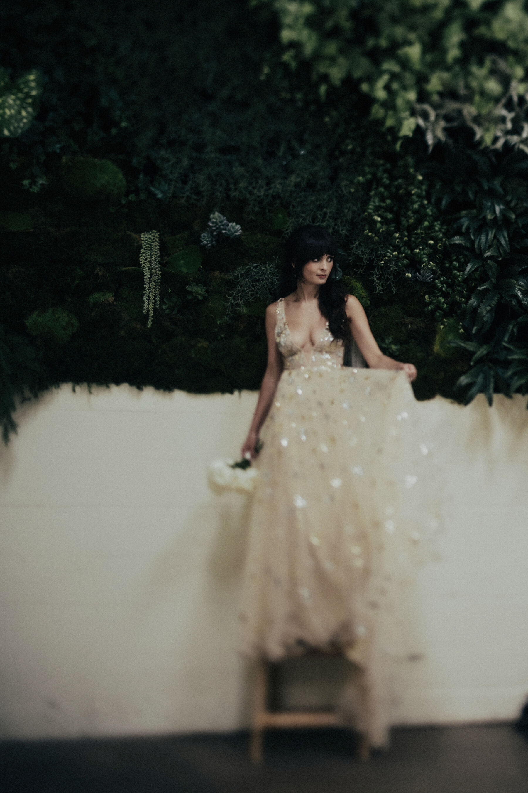 Long, dark haired brie wearing the Orion dress by Watters - a celestial inspired wedding dress. She carries a bouquet of white flower. The photograph is intentionally blurred in the bottom half.