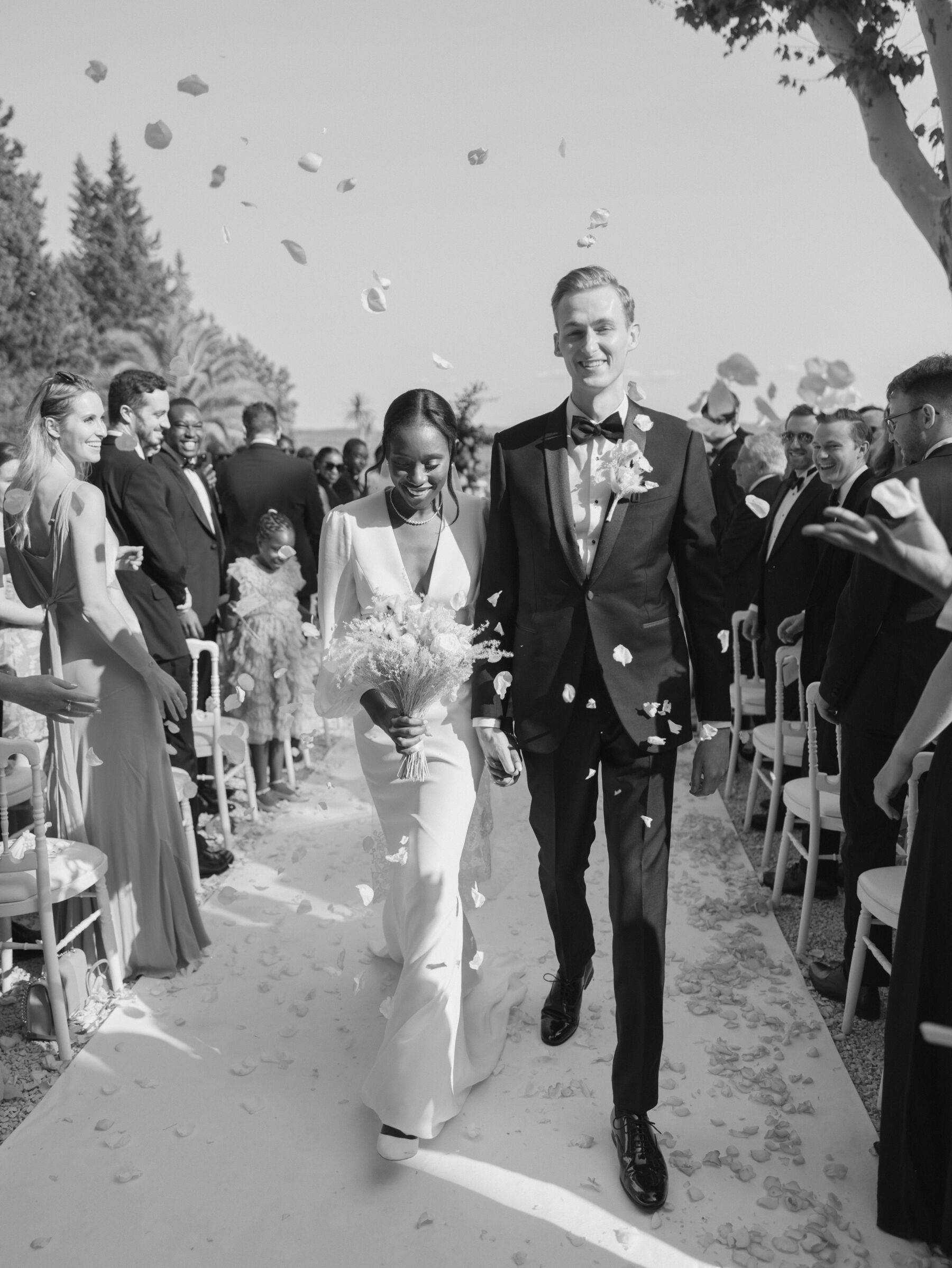 French Riviera Chic Wedding at Château Robernier, Provence, with Lawyer Chanel in Sassi Holford 26 Black bride wearing Sassi Holford and her groom walking through a confetti shower as they exit their outdoor wedding ceremony. Wedding at Chateau Robernier in Cotignac, Provence, France. Sébastien Boudot Photography.