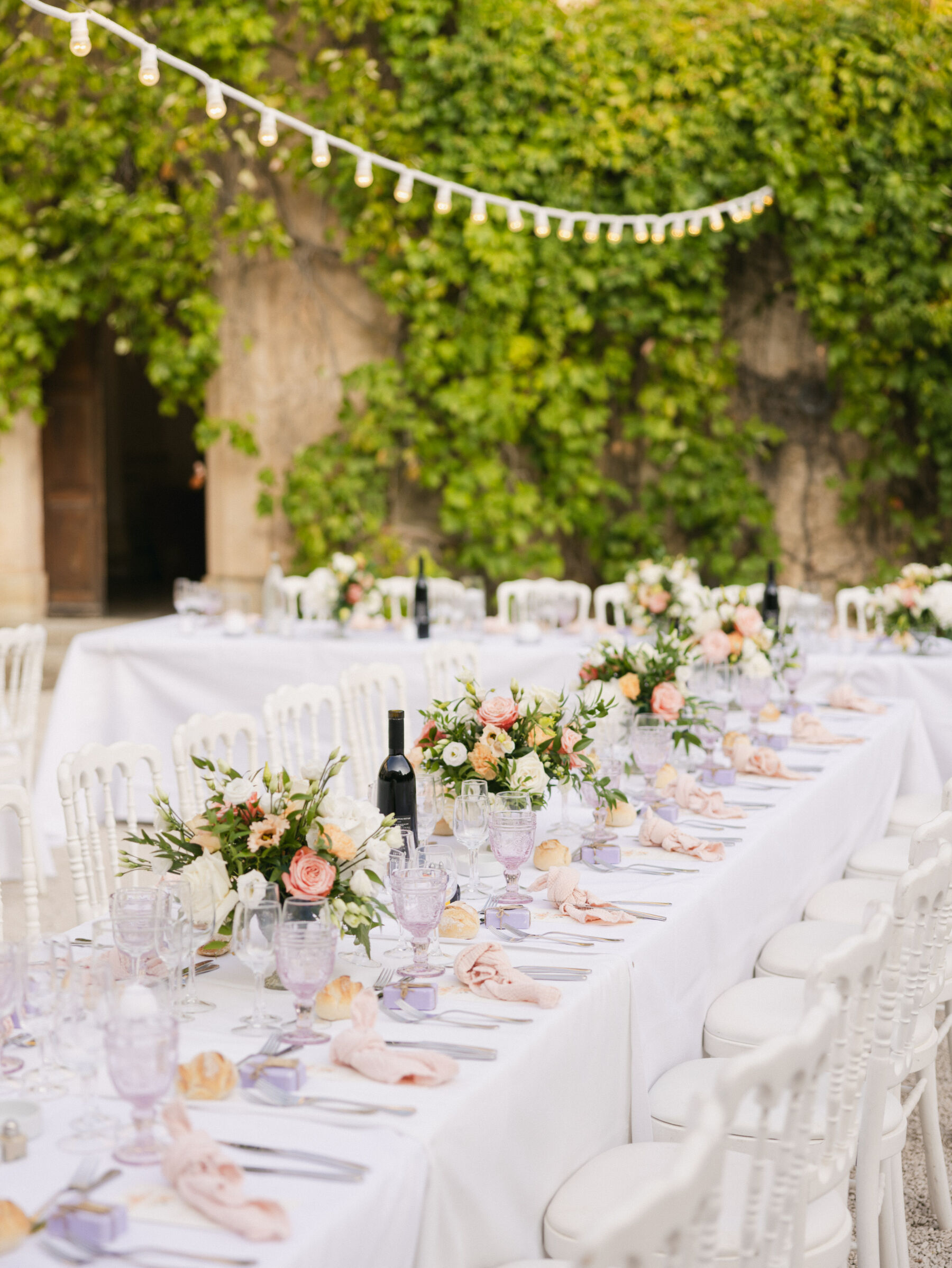 French Riviera Chic Wedding at Château Robernier, Provence, with Lawyer Chanel in Sassi Holford 33 Wedding reception setting at Chateau Robernier in Cotignac, Provence, France. Festoon lights hang above long tables covered in white table cloth with pink napkins and summer flowers as table centrepieces. White chairs.