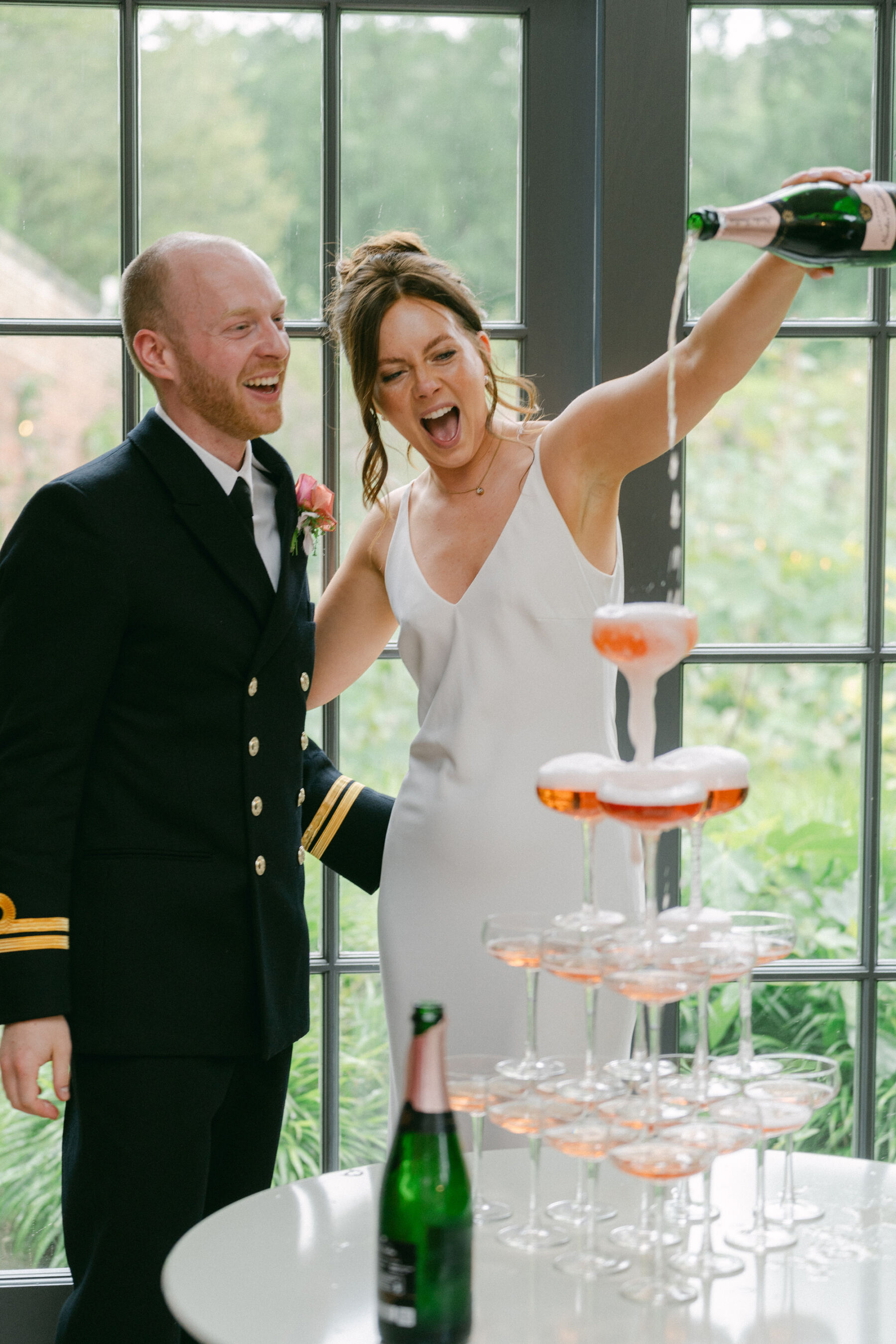 Bride excitedly pouring into the top glass of a champagne tower - her groom in navy military uniform smiling nearby.