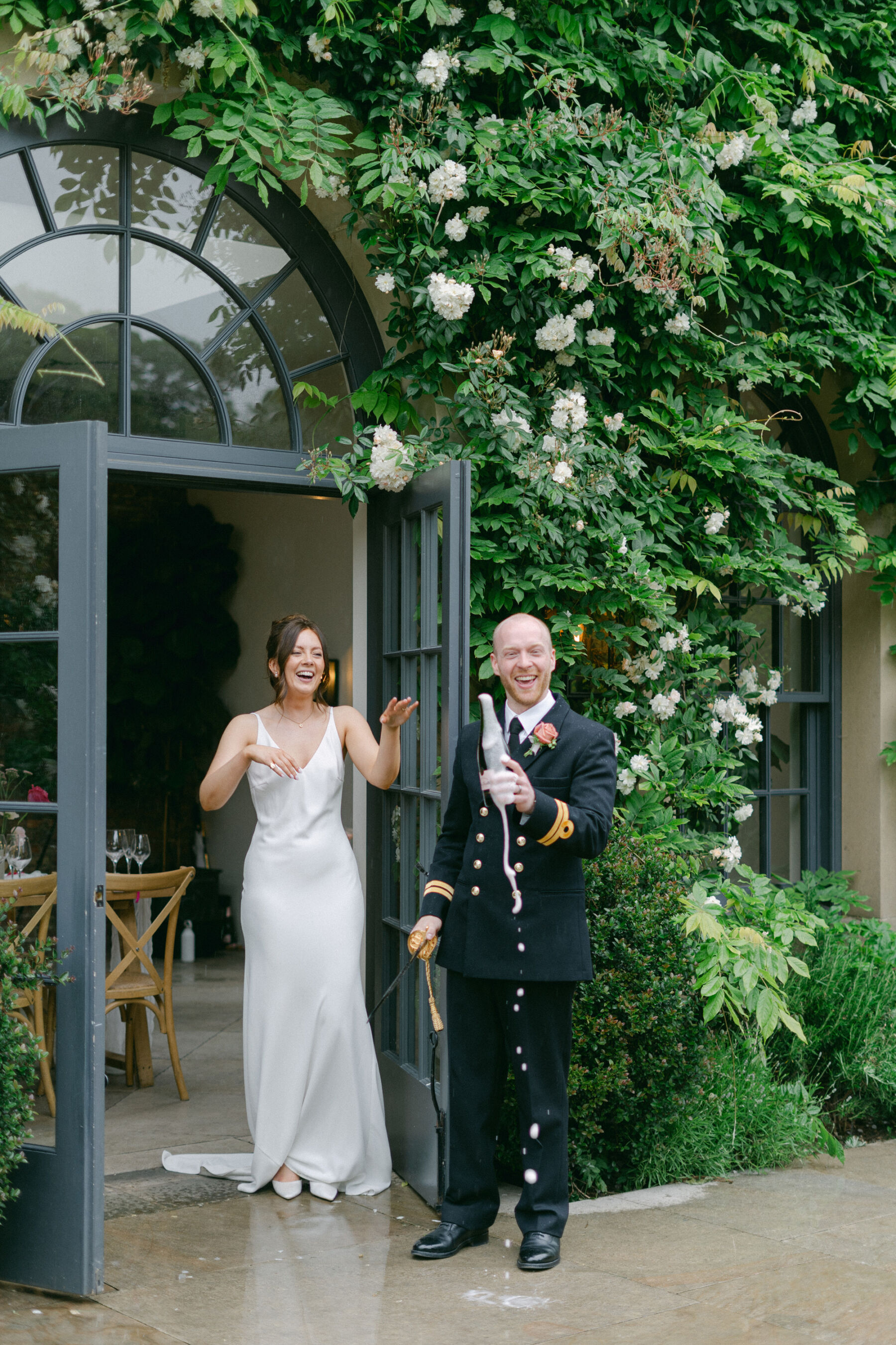 Groom in navy military uniform popping the champagne outside Middleton Lodge with his bride in a simple wedding dress standing nearby.