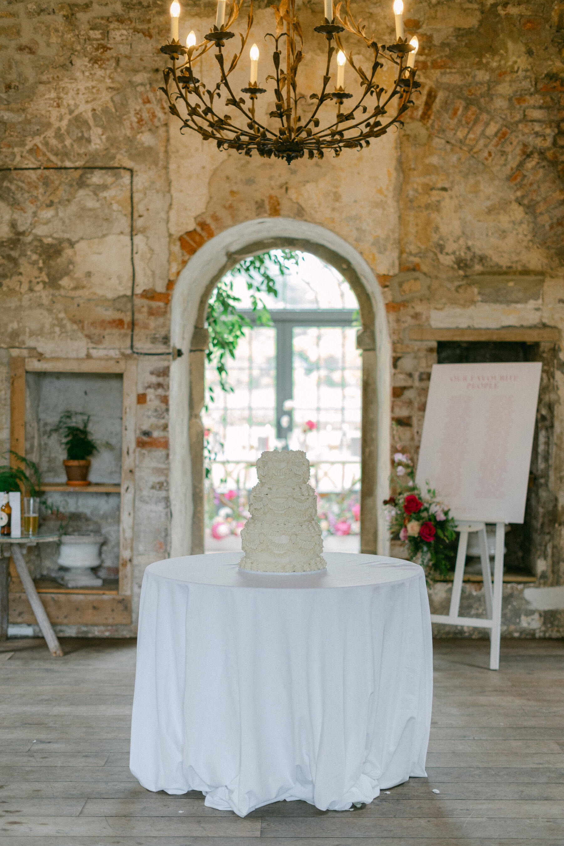 Wedding cake standing on a small round table draped in white table cloth. Above the table is a cast iron chandelier with candles. Rustic wedding reception space at Middleton Lodge wedding.