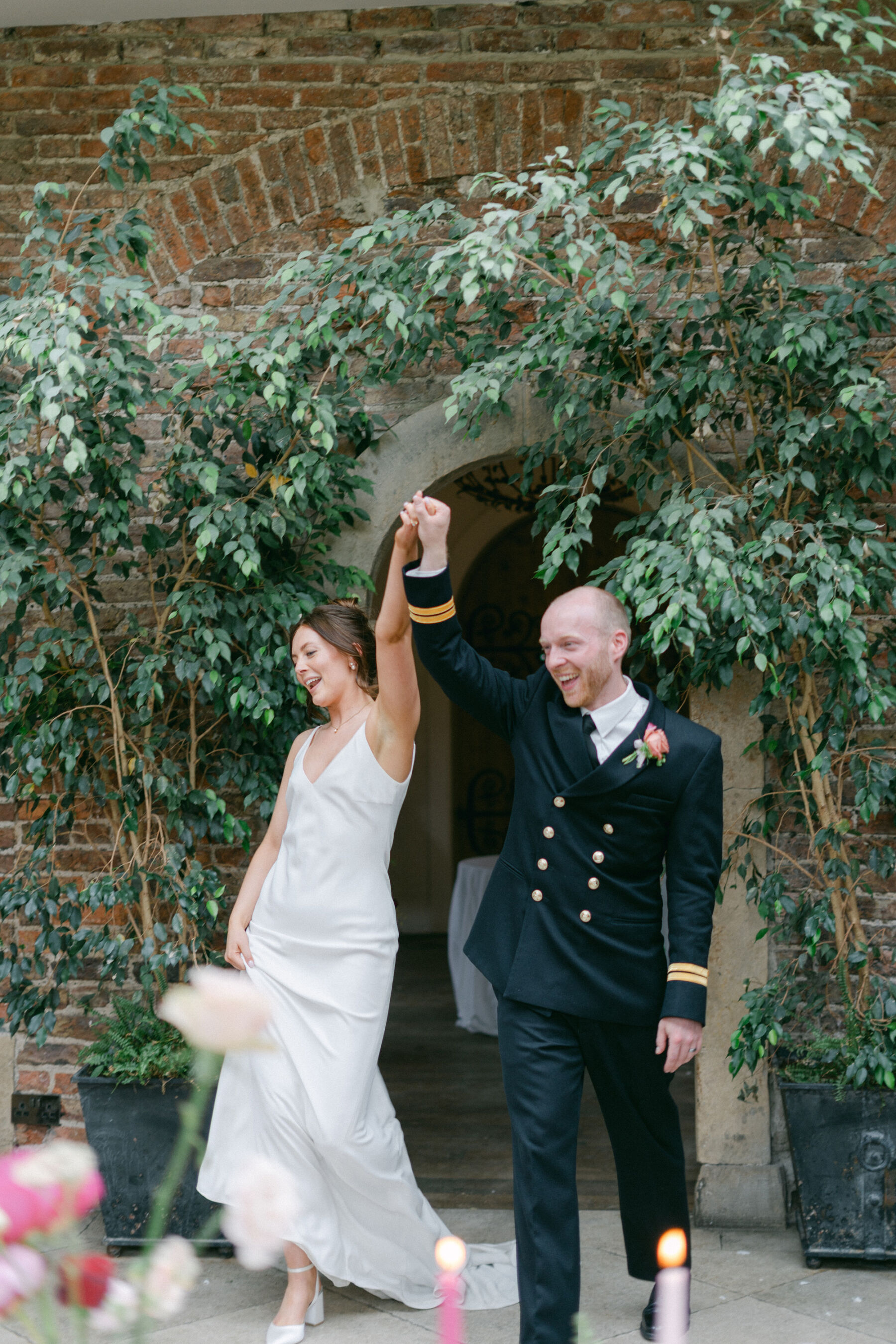Bride and groom entering the Fig Room at Middleton Lodge for their wedding reception, they are raising their held hands in the air. The bride wears a simple wedding dress and the groom is in Royal Navy military uniform.