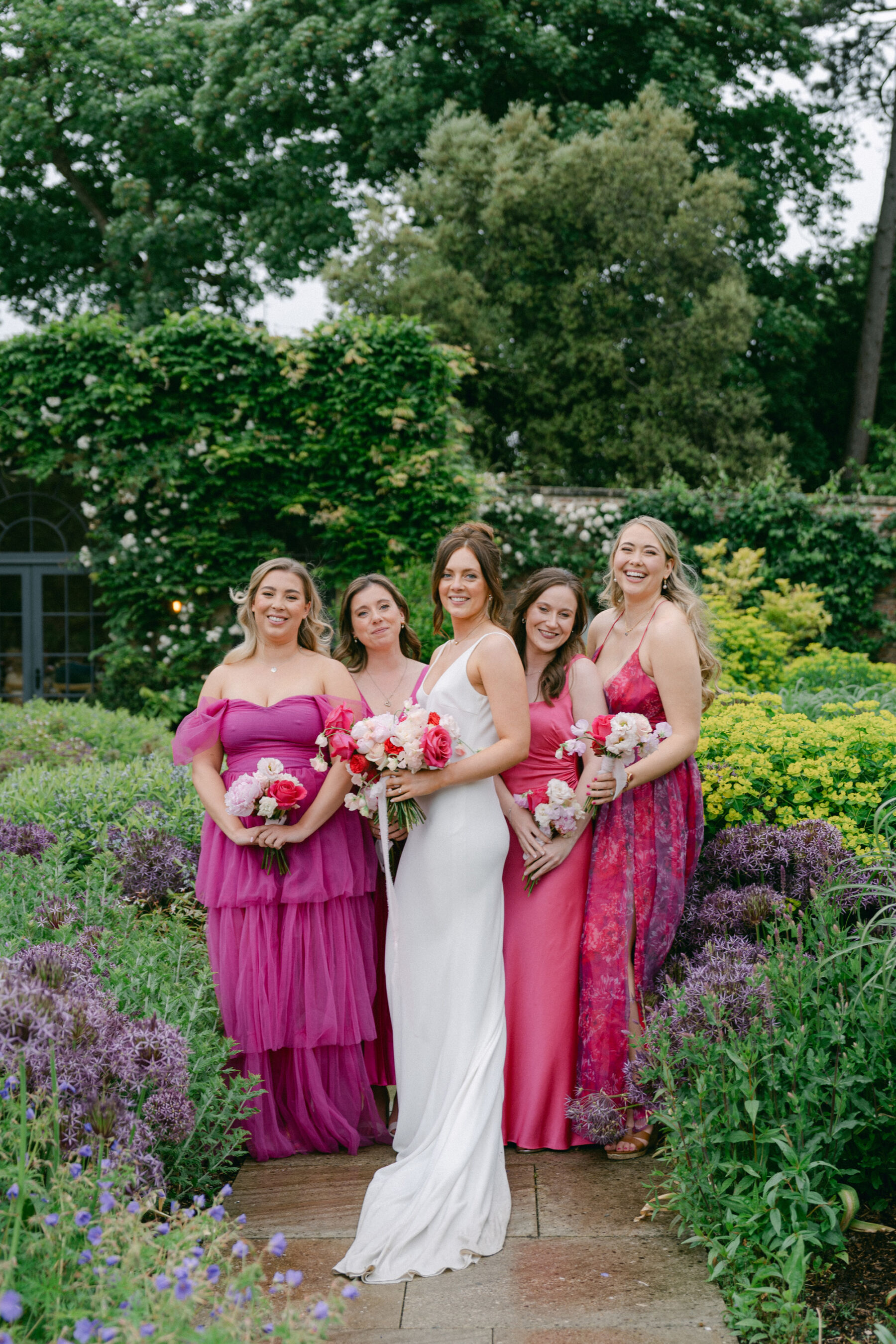 Bride in a simple minimalist wedding dress surrounded by bridesmaids in bright pink dresses at Middleton Lodge. They all carry bouquets of roses in various shades of pink.