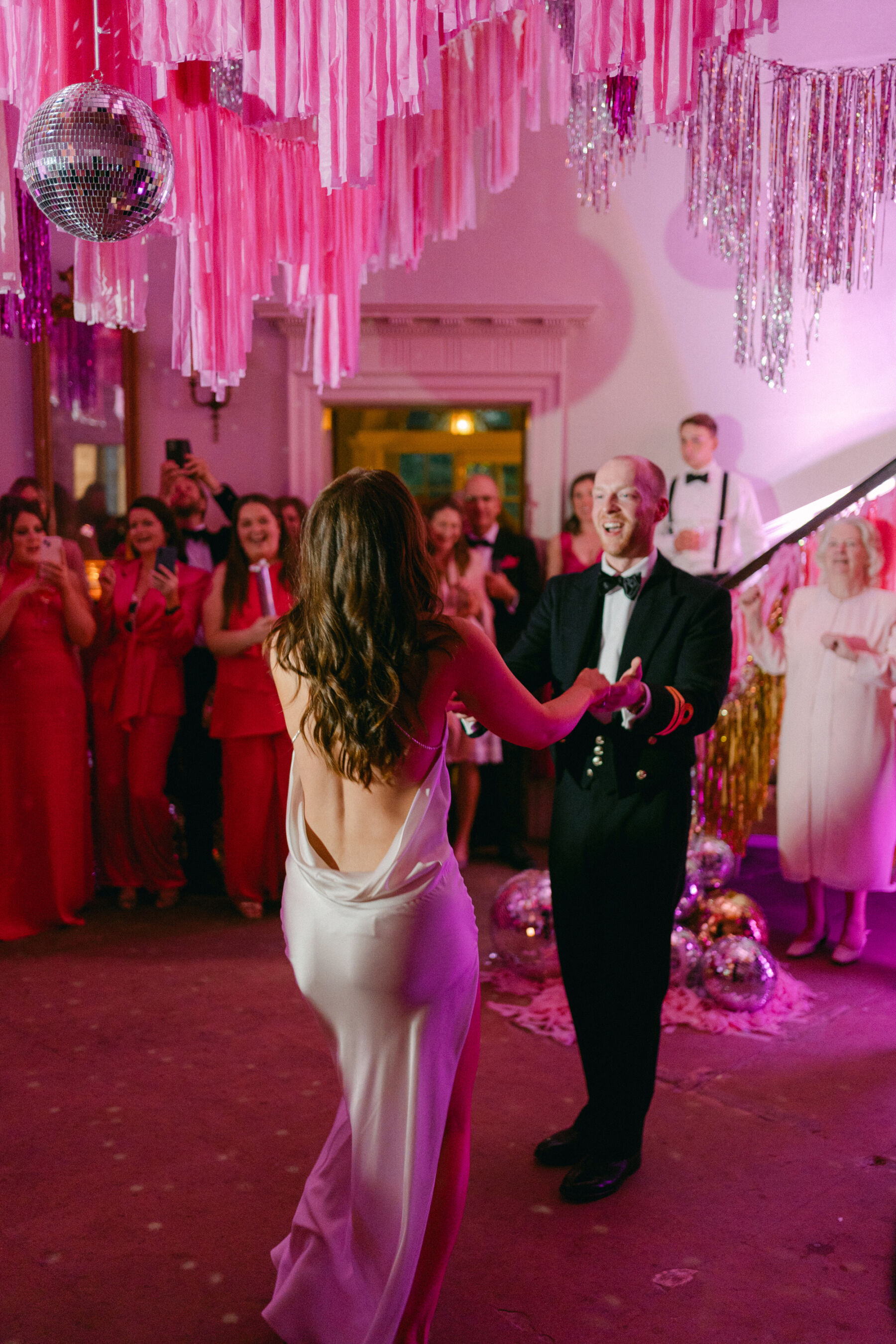 Bride in a backless wedding dress and loose hair dancing with her husband in navy military uniform on a dance floor at Middleton Lodge that is bathing in pink light. Above them pink and silver streams/tassels hang above them. 