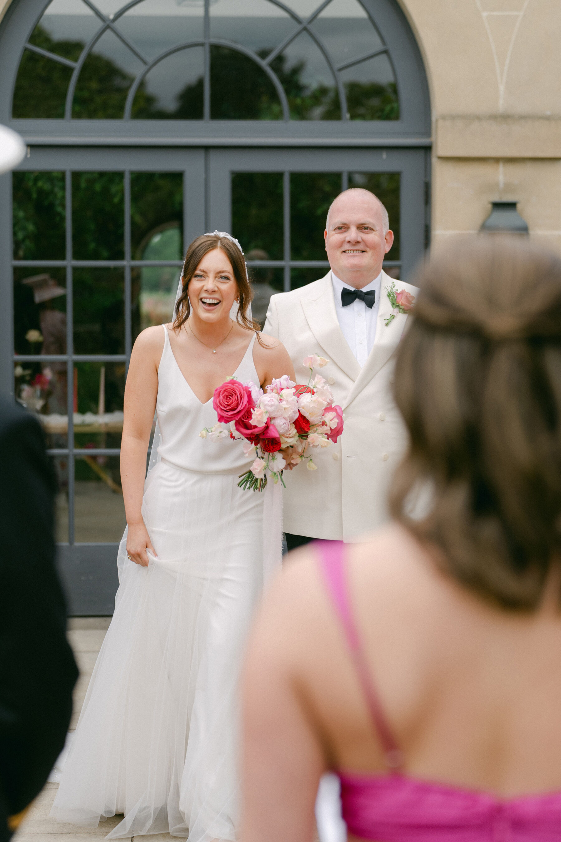 Bride being accompanied down the aisle by her father in a white tuxedo. She wears a minimalist wedding dress and carrie a bouquet of pink roses.