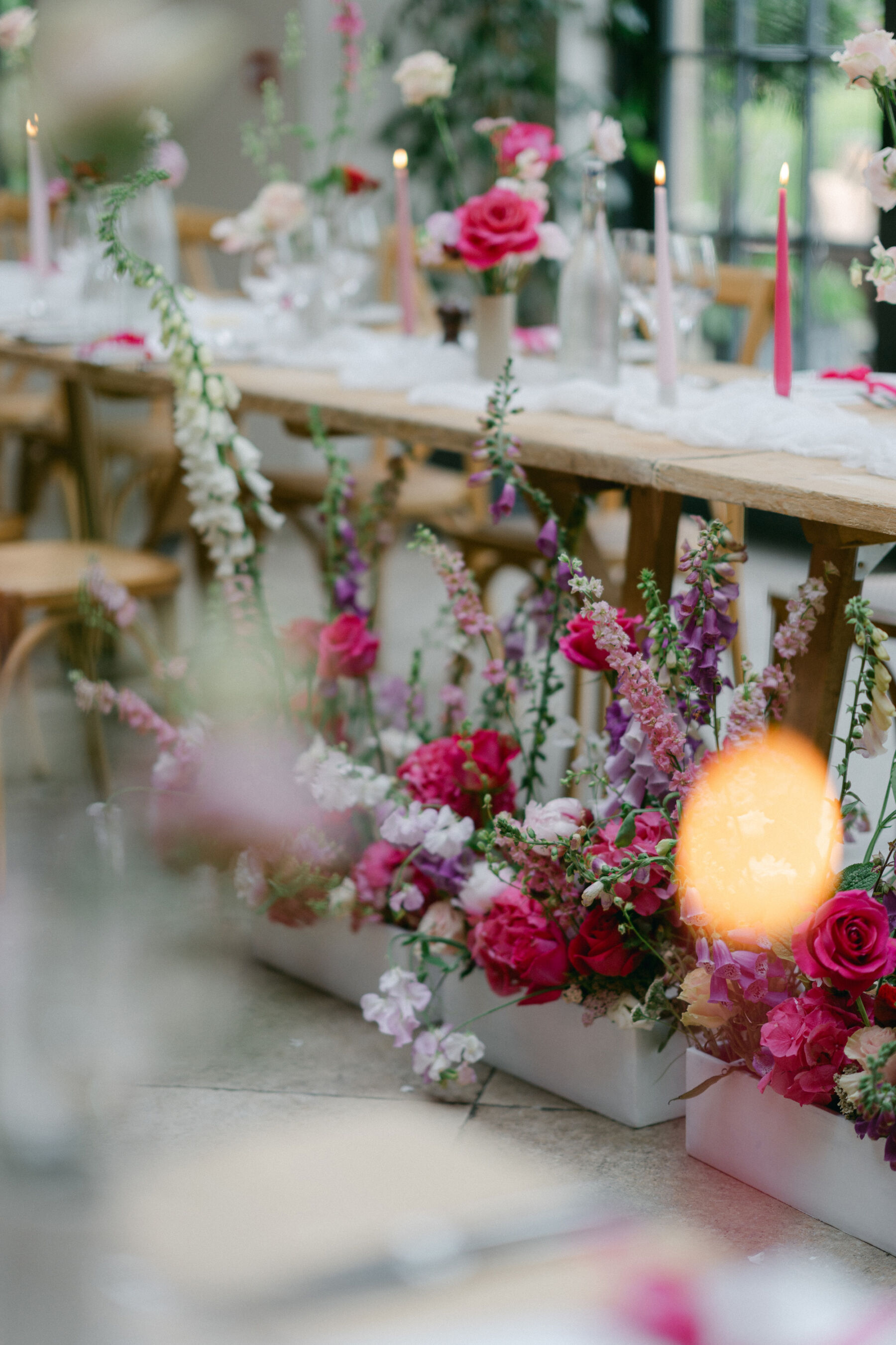 Pink wedding flowers in white trays rested on the floor next to long wooden trestle tables with pink taper candles. 