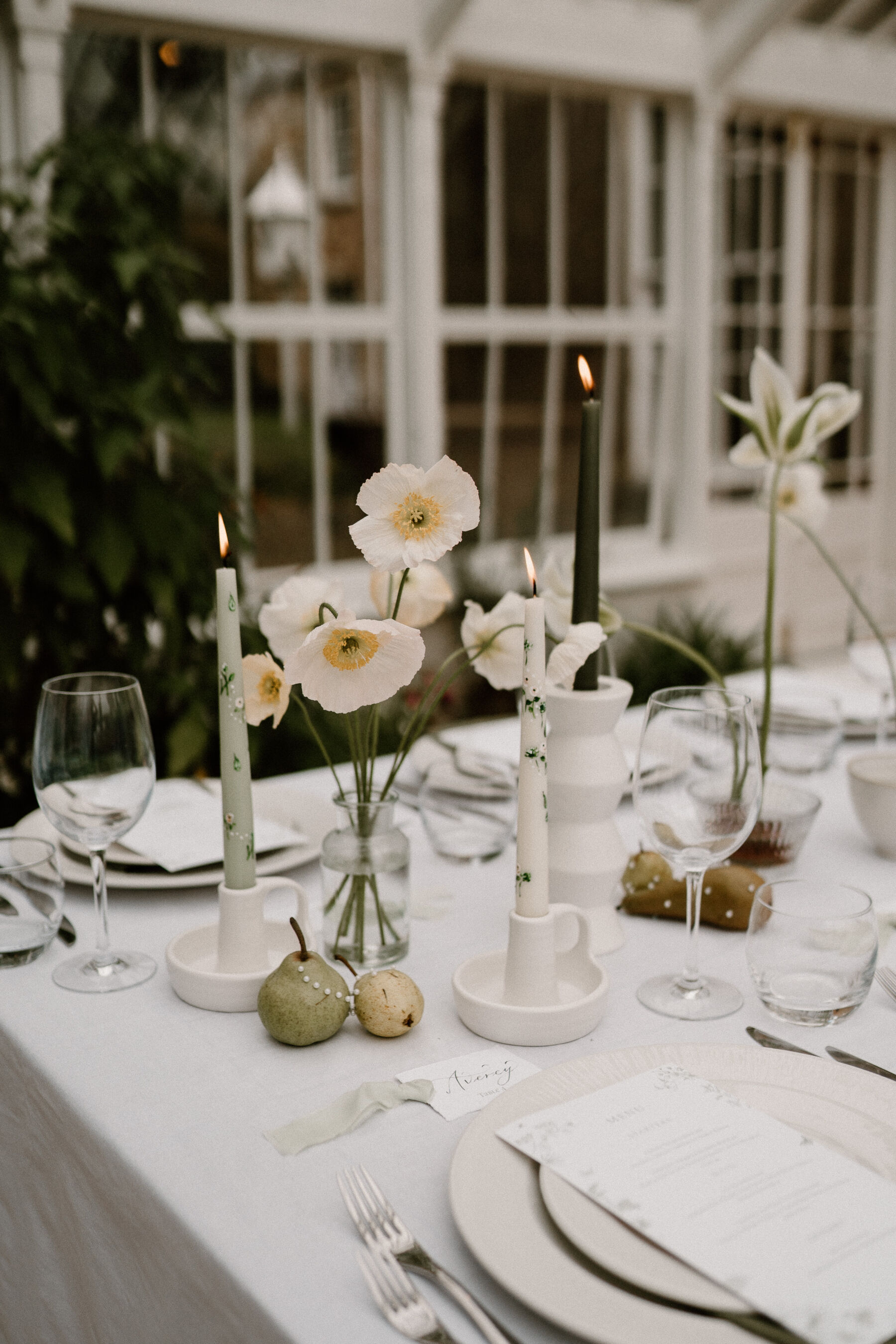 Elegant sage green modern spring wedding table with hand-painted taper candles, minimalist spring flowers in glass bud vases and pears decorated with pearls. 