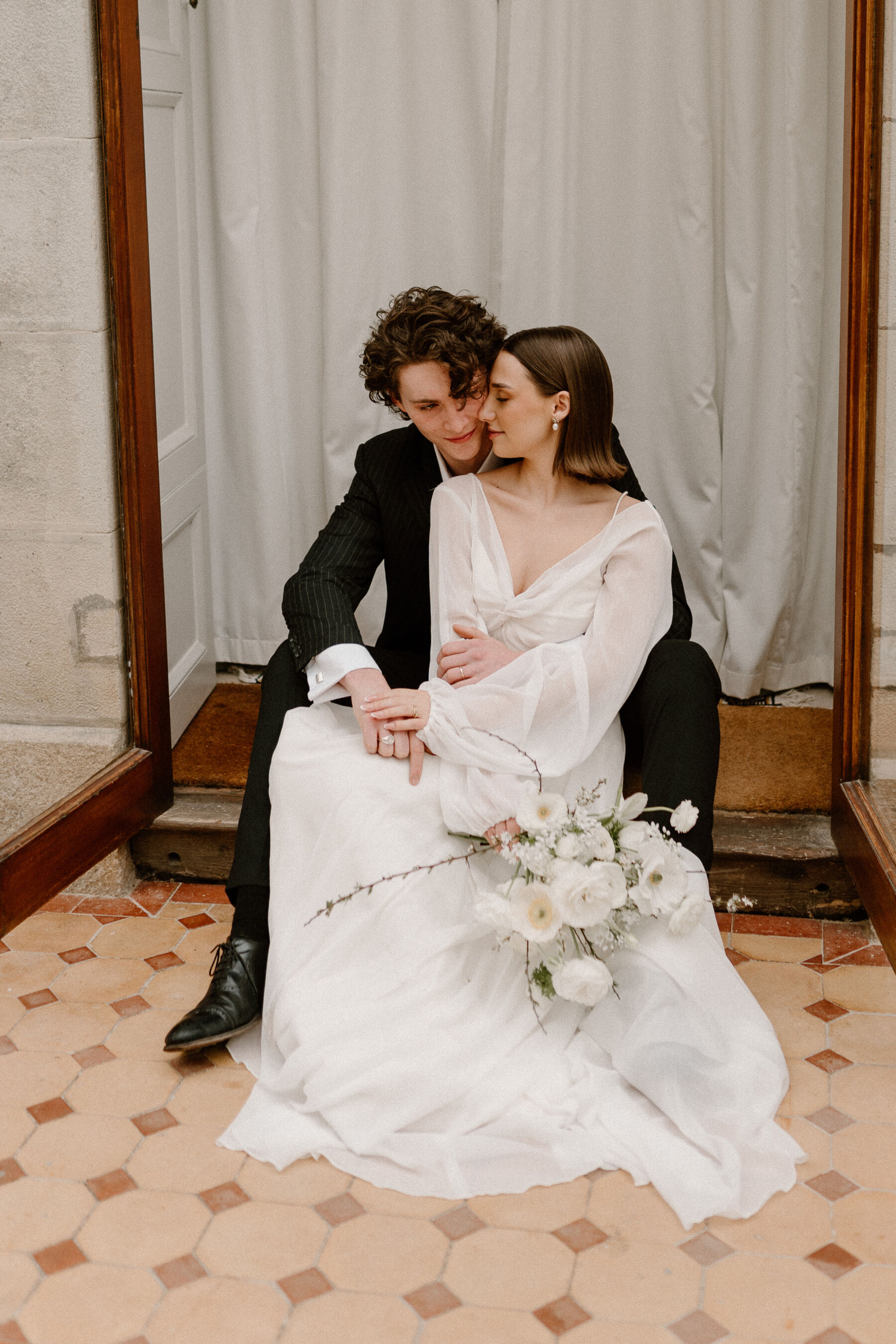 Young model bride and groom embracing each other whilst seated on a step. The bride wears an E&W Couture dress and carries an elegant spring wedding bouquet of white flowers and foraged twigs.