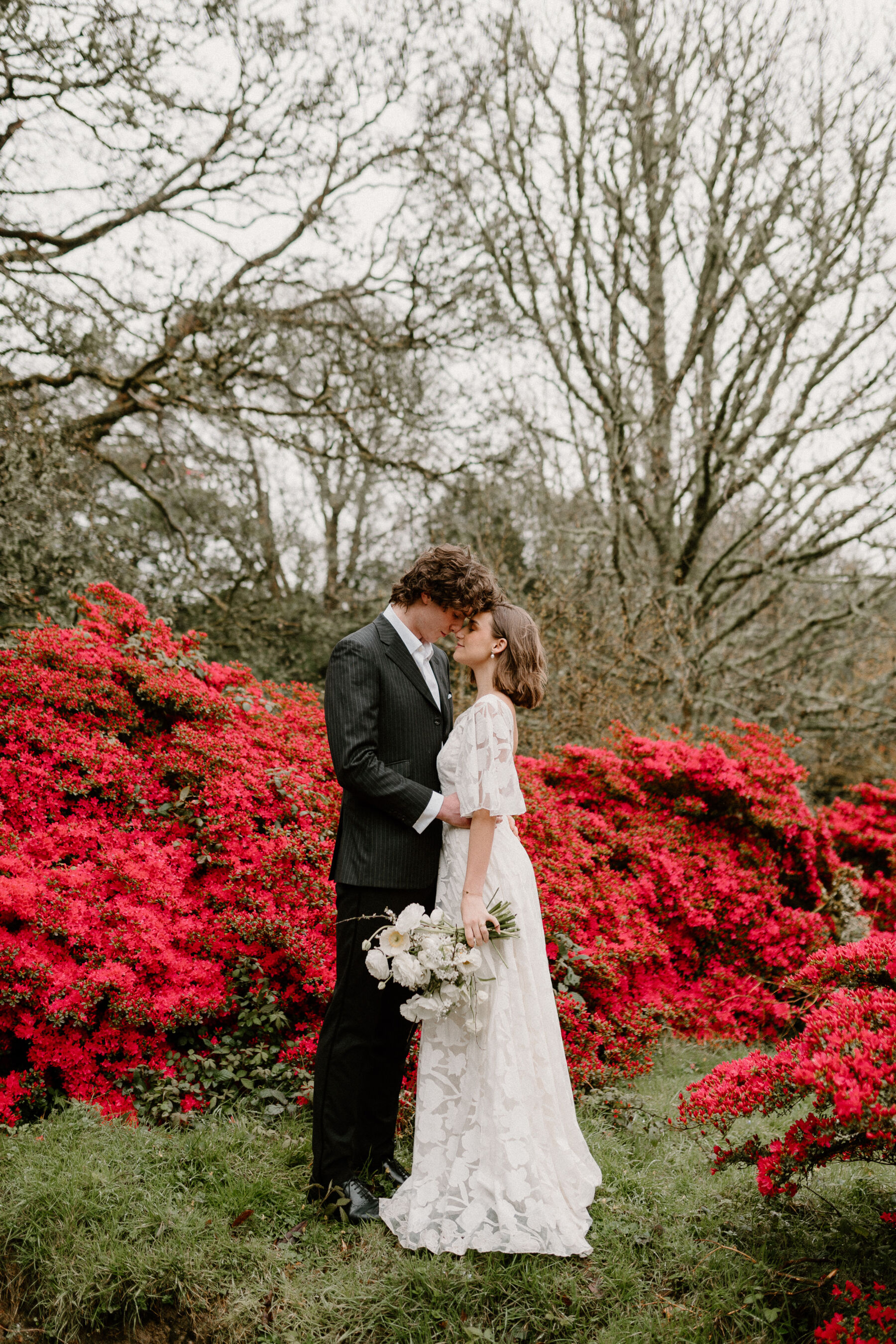 Bride and groom standing by a bright red colour bush. 