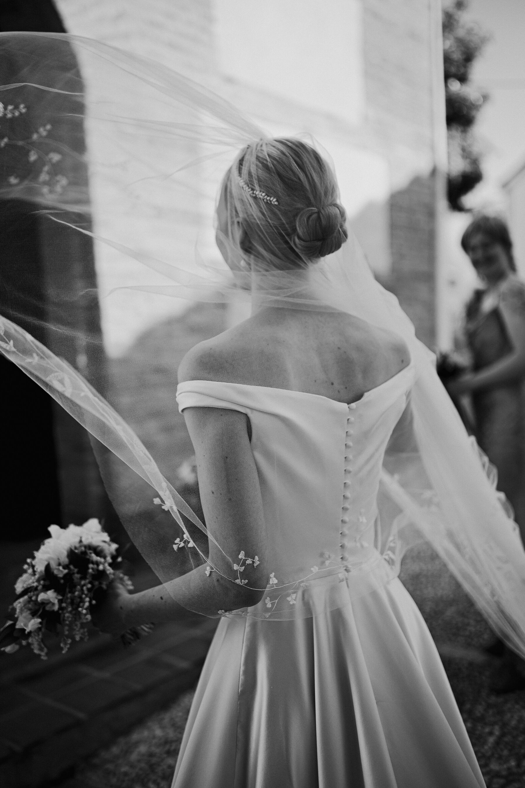 Alexandra Wore Phillipa Lepley For Her Romantic Destination Wedding in Spain 33 Black and white photograph from behind of elegant blonde bride with a simple bun wearing a Phillipa Lepley dress and embroidered veil. She carries a small bridal bouquet.