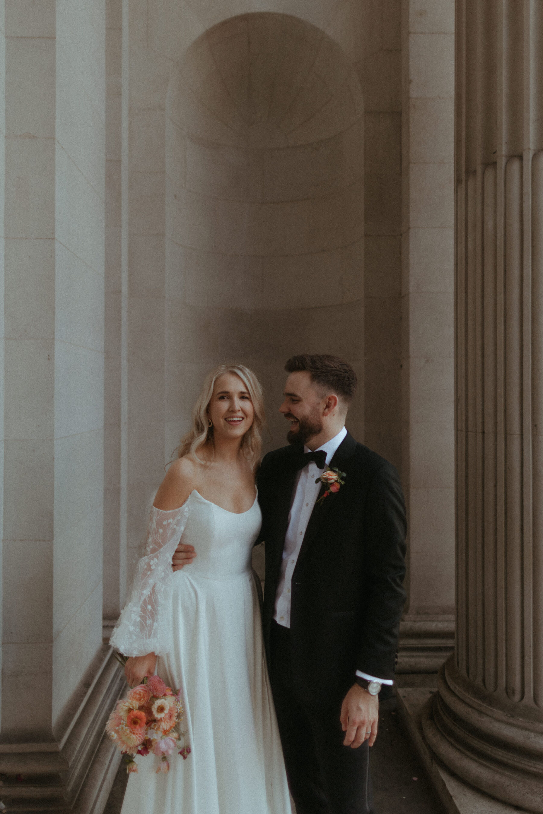 Georgia in Truvelle for her Modern London Wedding at The Depot in Islington 18 Bride wearing a Truvelle wedding dress with detachable sleeves being hugged around the waist by her groom in black tie.