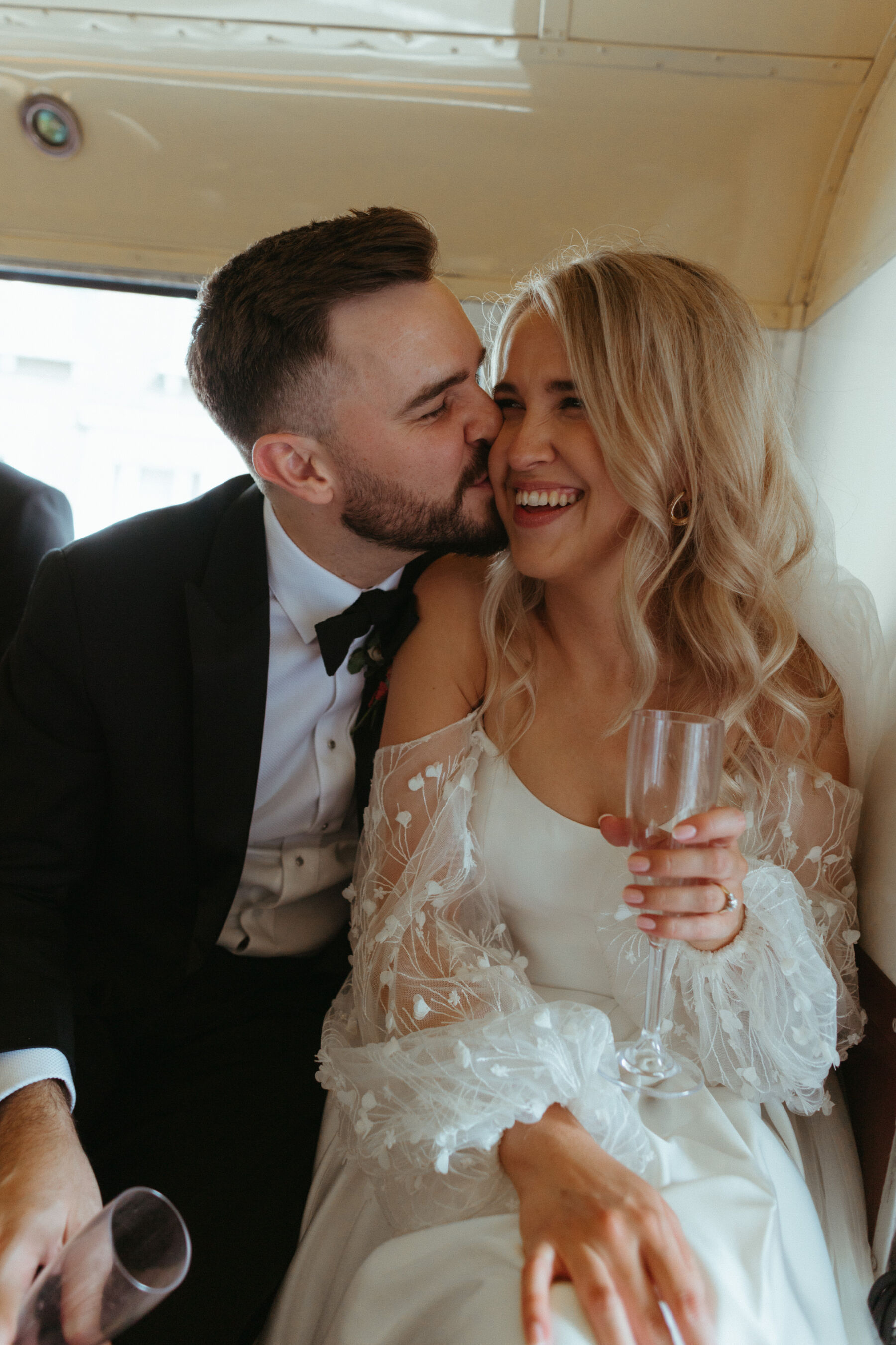 Georgia in Truvelle for her Modern London Wedding at The Depot in Islington 17 Bride and groom smooching inside their wedding day transport with glasses of wine. The bride has a very pale pink manicure, modern gold hoop earrings, long and loose wavy blond hair and is wearing a strapless Truvelle wedding dress with detachable sleeves. Groom in black tie.