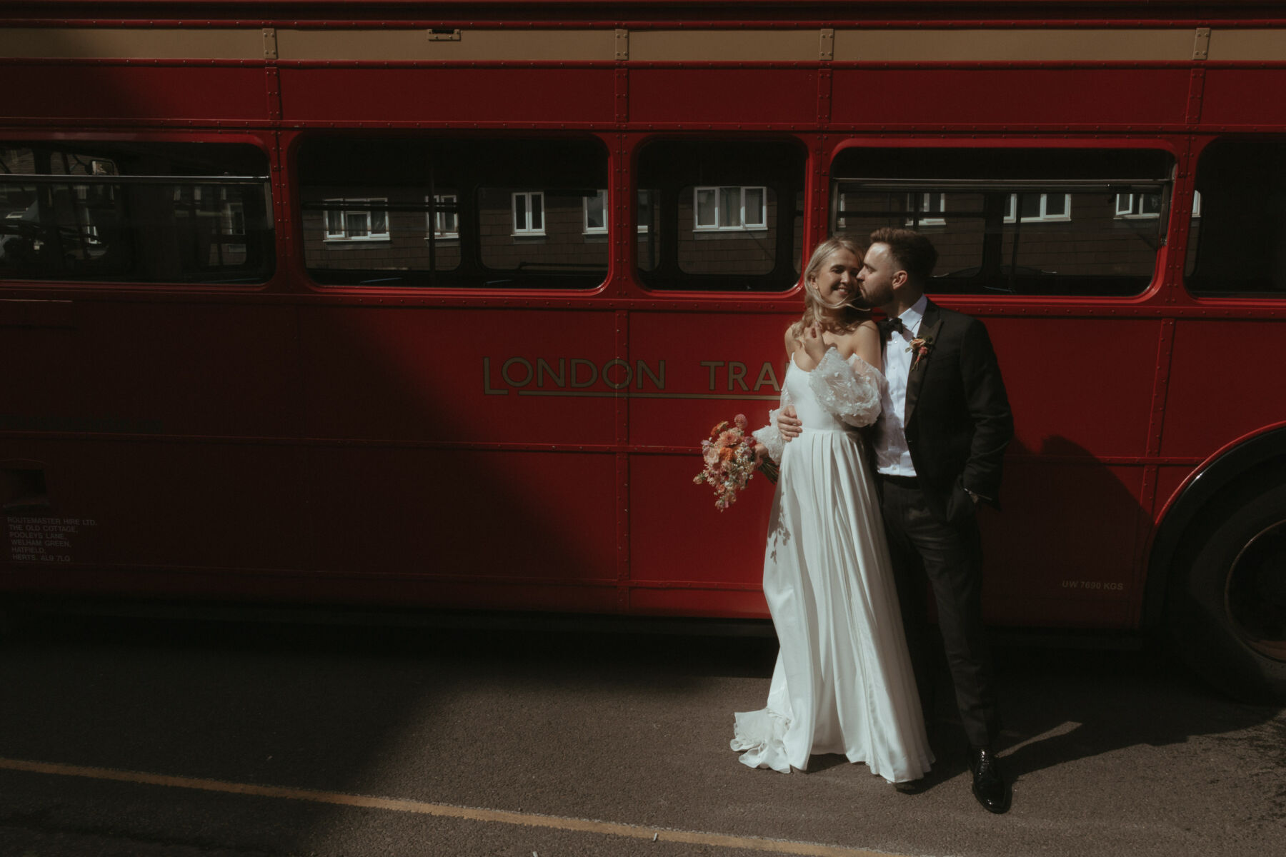 Georgia in Truvelle for her Modern London Wedding at The Depot in Islington 21 Bride and groom standing beside their wedding day red routemaster bus. The bride wears Truvelle and the groom is in black tie